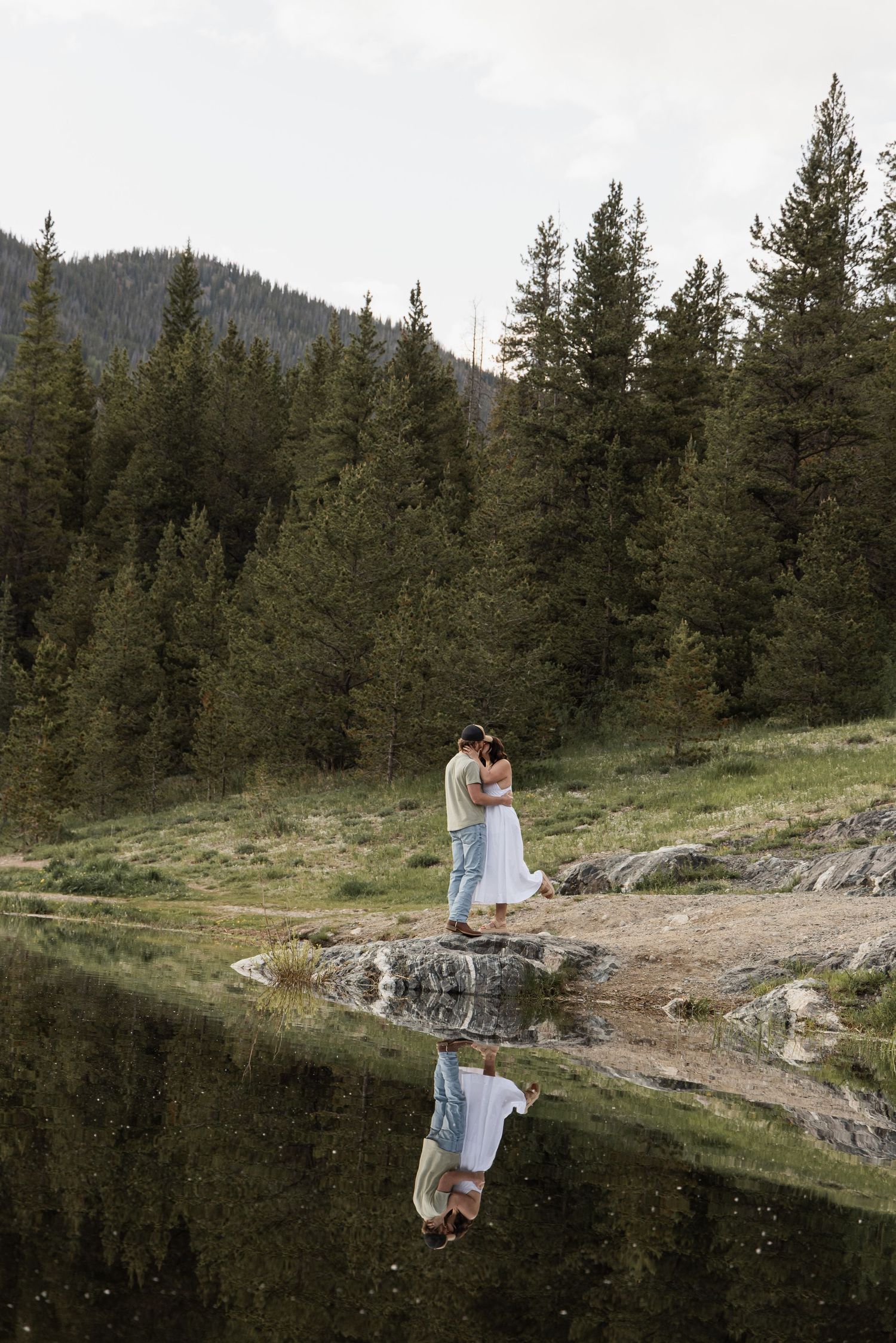 A couple embraces beside a tranquil mountain lake reflecting pine trees and forest landscape in the background.
