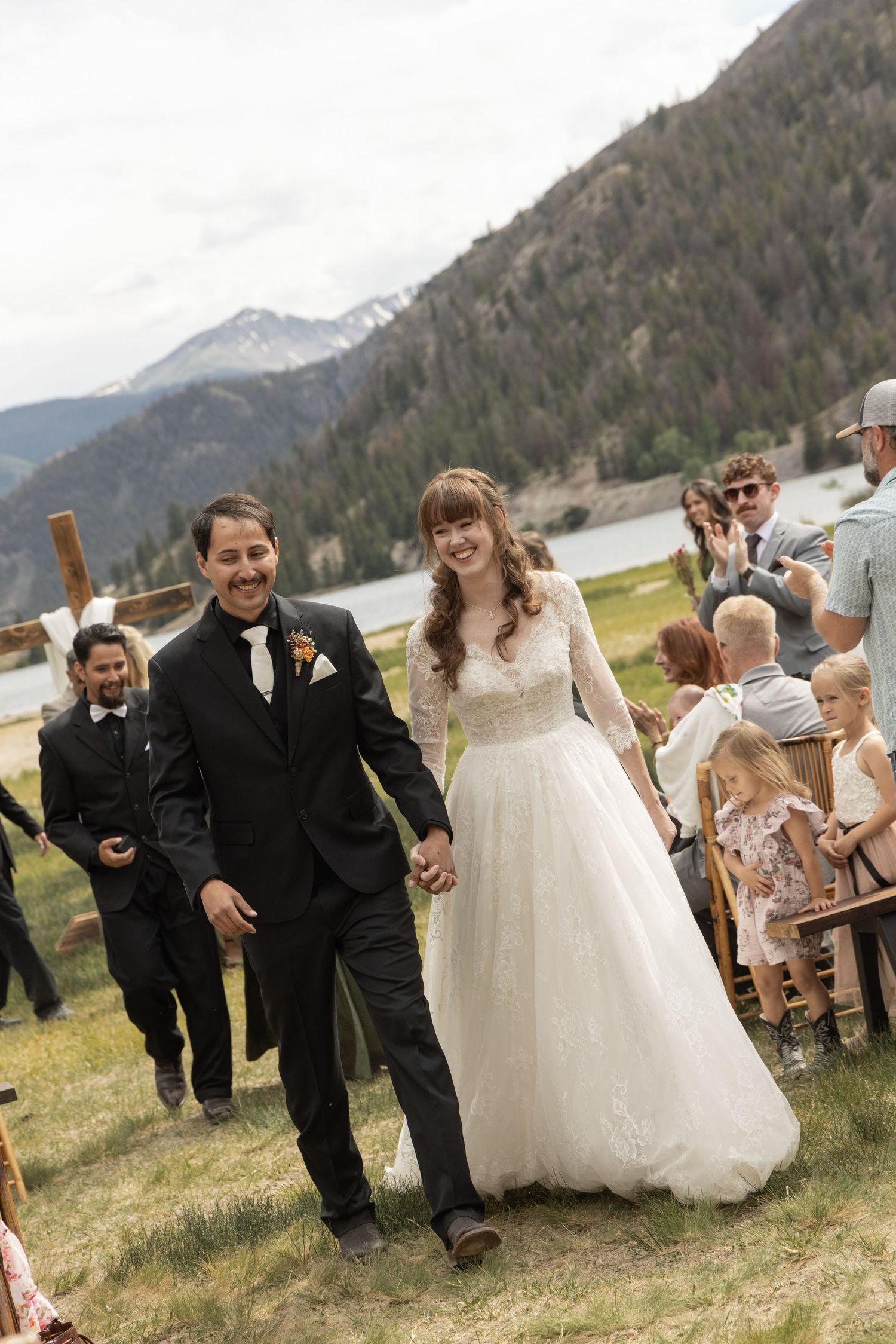 Bride and groom walk down the aisle at an outdoor mountain wedding ceremony, smiling joyfully with guests and scenic peaks behind.