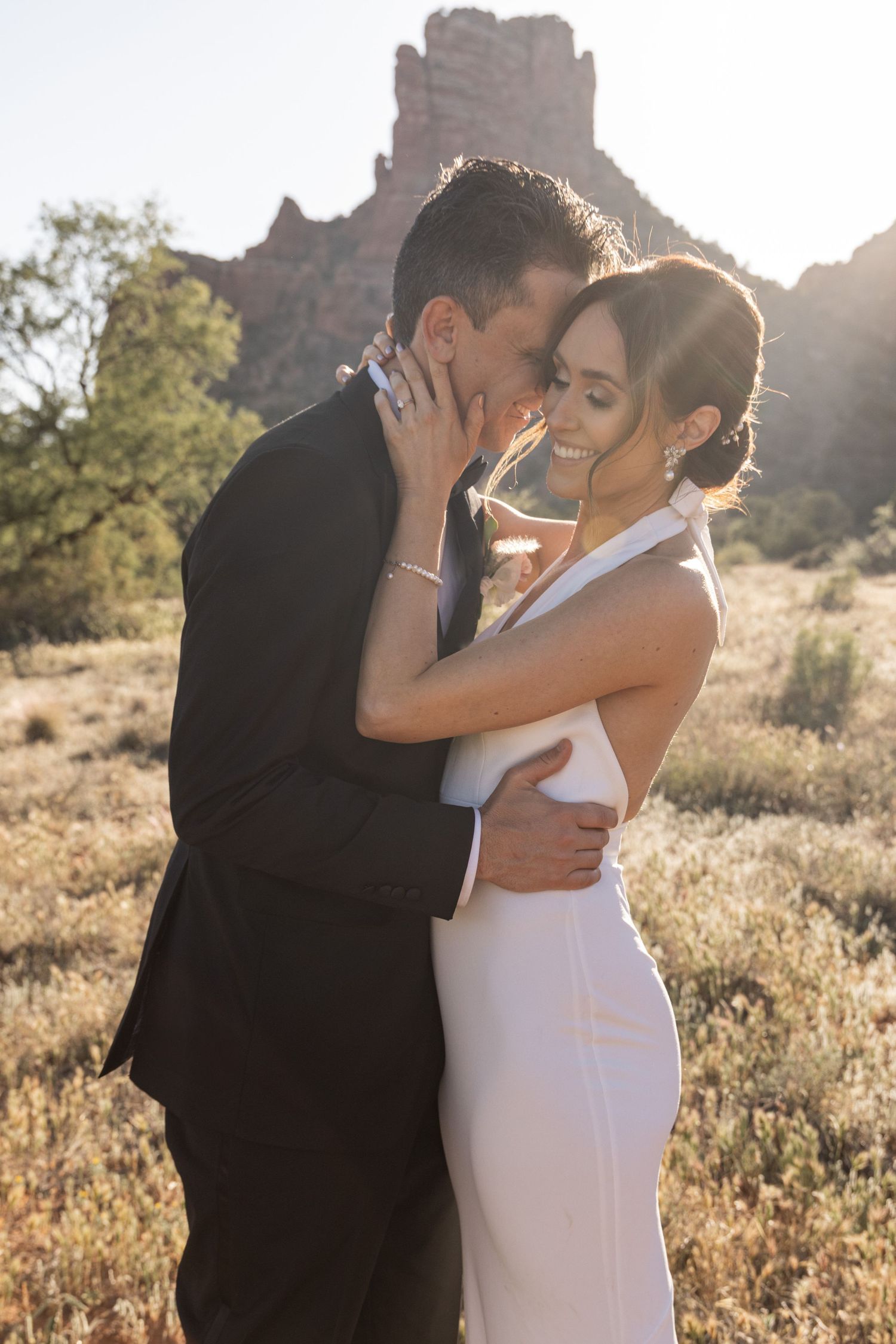 A romantic couple embraces in golden sunlight among desert brush and rock formations during a sunset photo session.