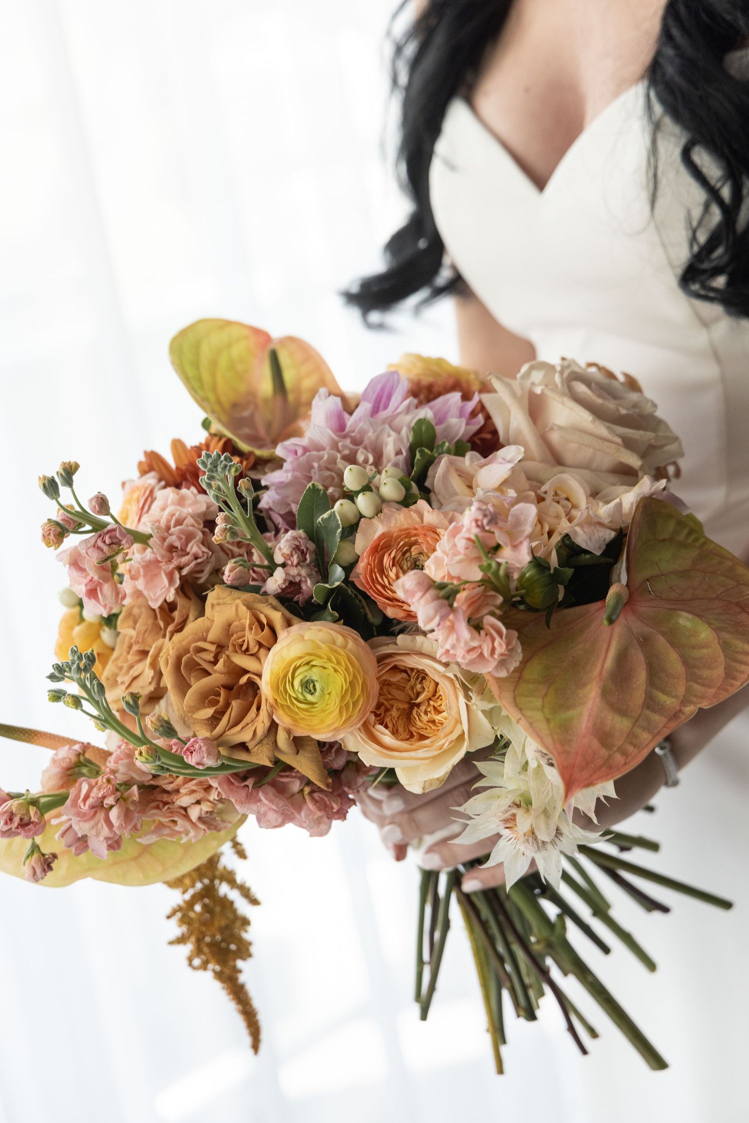 Bride in white dress holding lush autumn wedding bouquet with peach roses, ranunculus, and anthurium blooms.