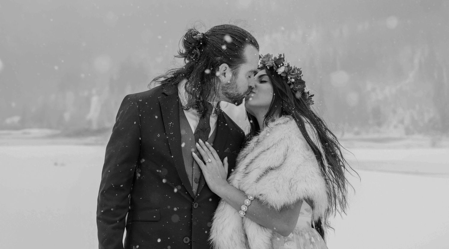 Couple sharing a romantic kiss in falling snow, groom in suit, bride in fur wrap and floral crown, black and white.