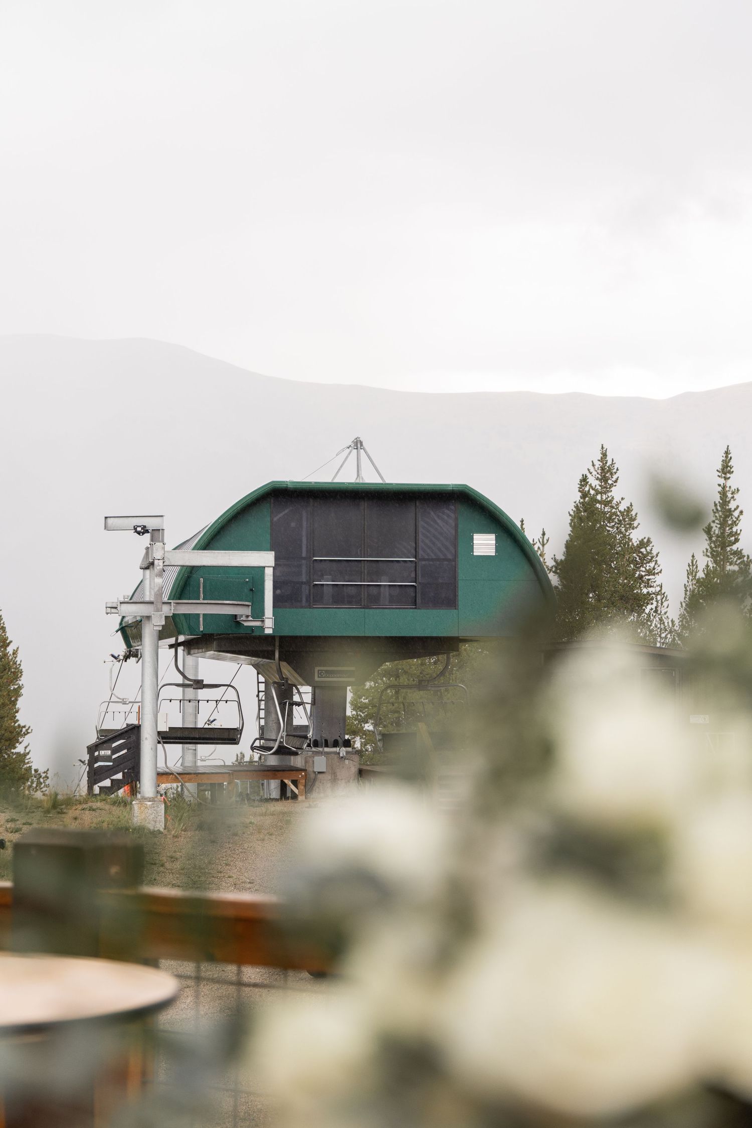 Green ski lift terminal building on a mountain top surrounded by pine trees and misty mountain backdrop.