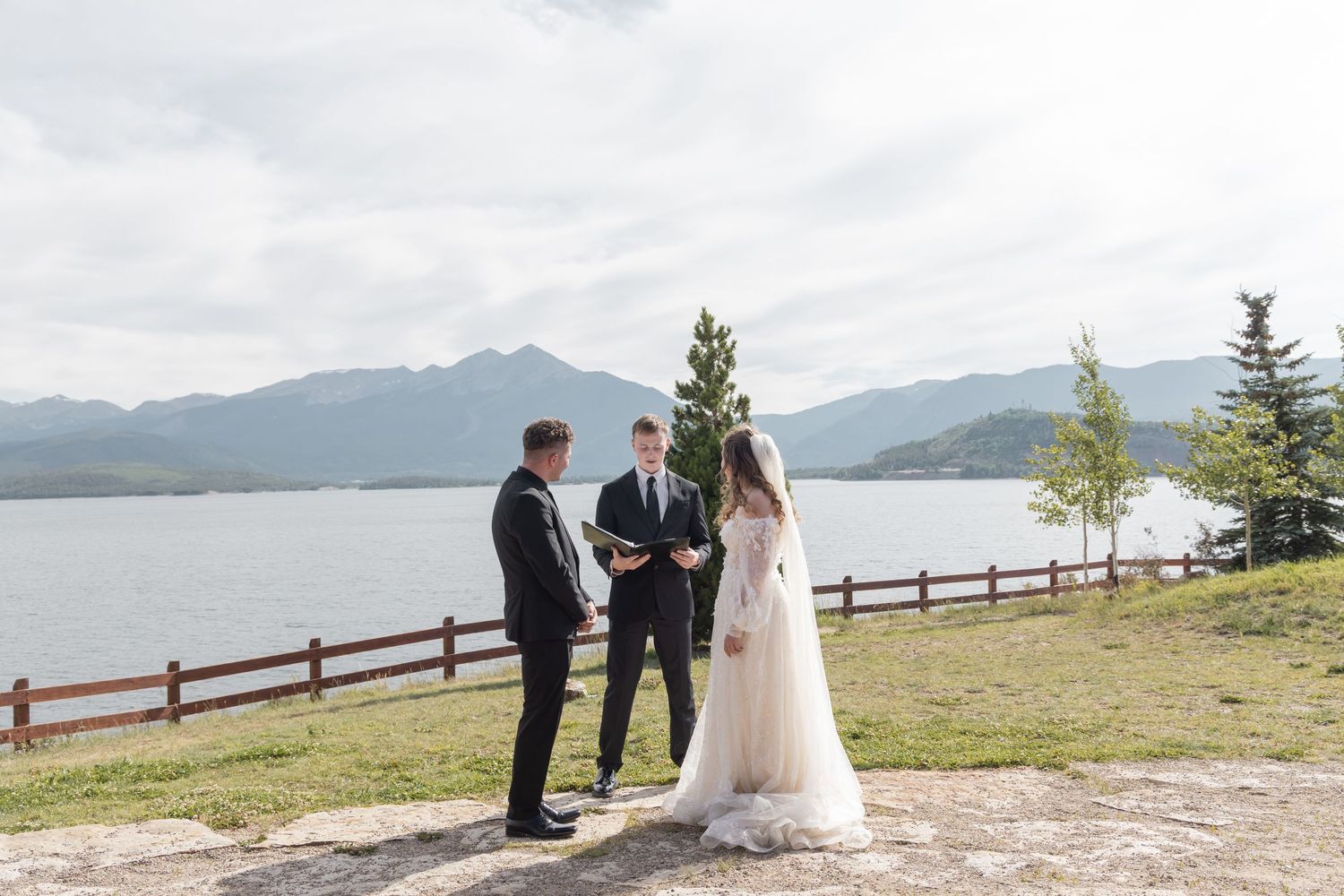 Outdoor wedding ceremony by a mountain lake with bride, groom, and officiant standing on a scenic overlook.