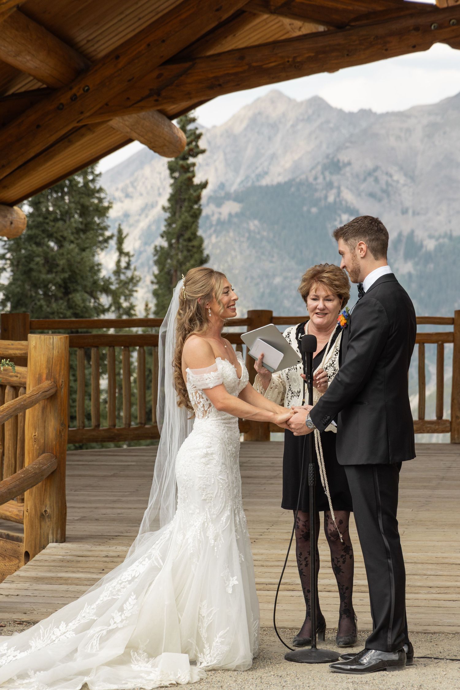Bride and groom exchange vows with officiant on a rustic mountain lodge deck with scenic peaks in the background.