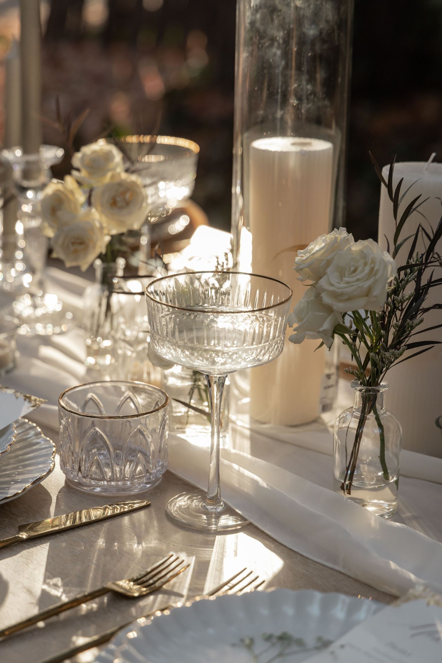 Elegant wedding table setting with crystal glassware, gold cutlery, white roses, and glowing candles in warm sunlight.