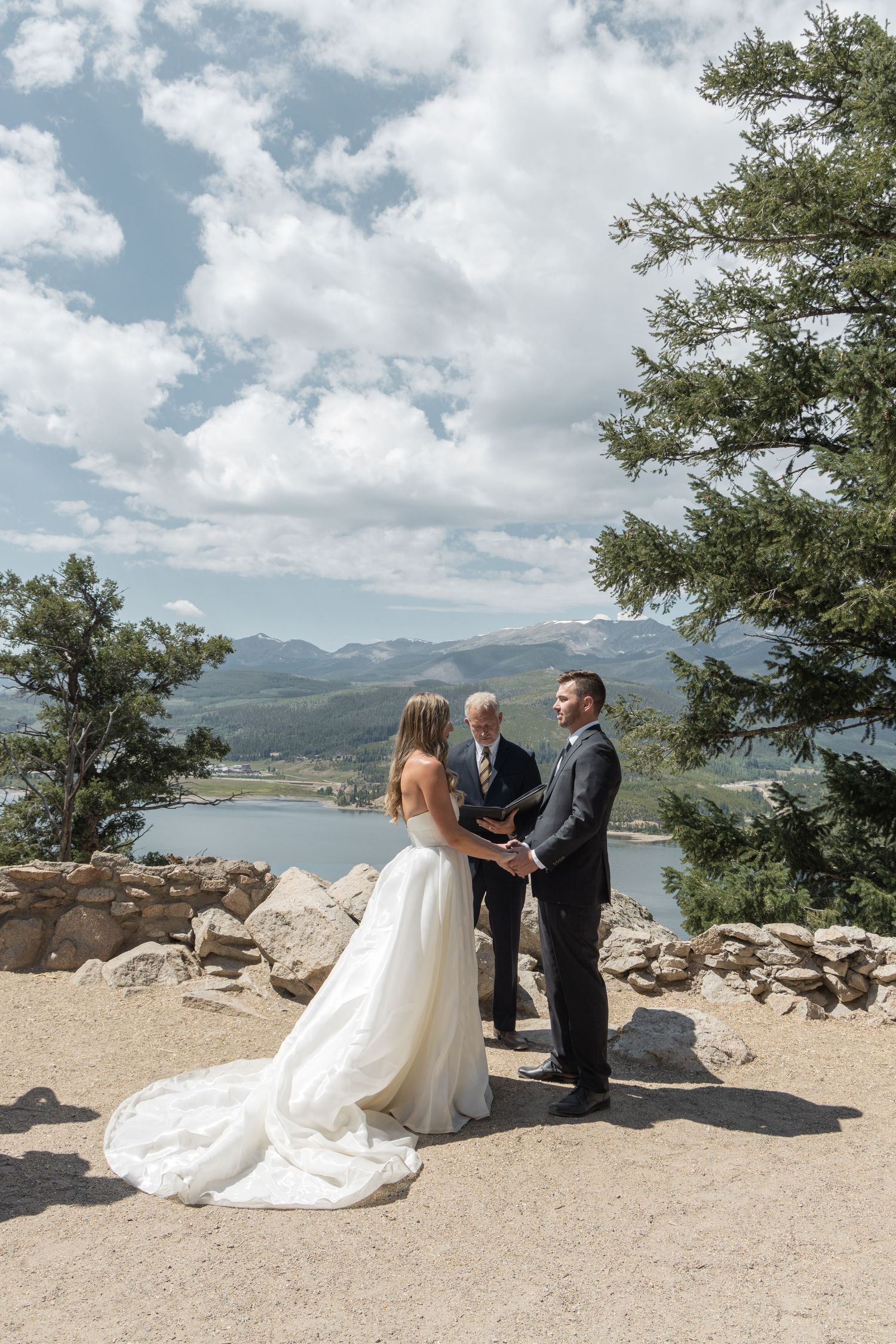 Couple exchanges vows at scenic mountain overlook wedding ceremony with lake and Rocky Mountain views under partly cloudy sky.