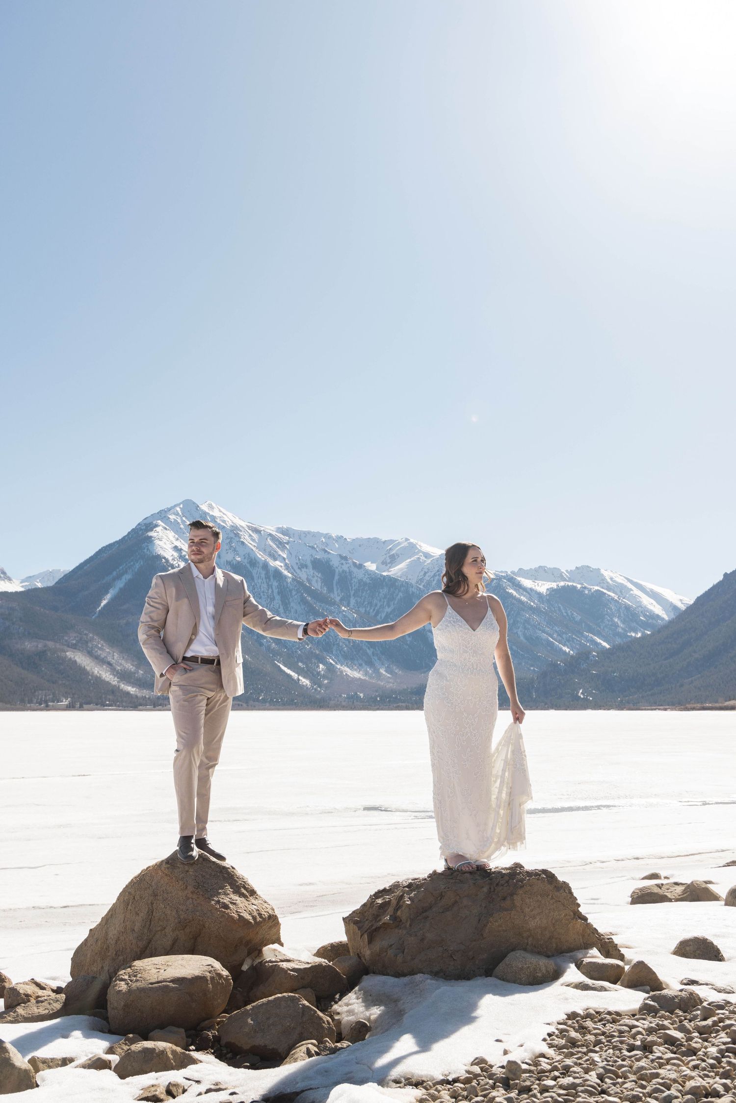Couple in wedding attire stand on snow-covered rocks with majestic snow-capped mountains in the background.