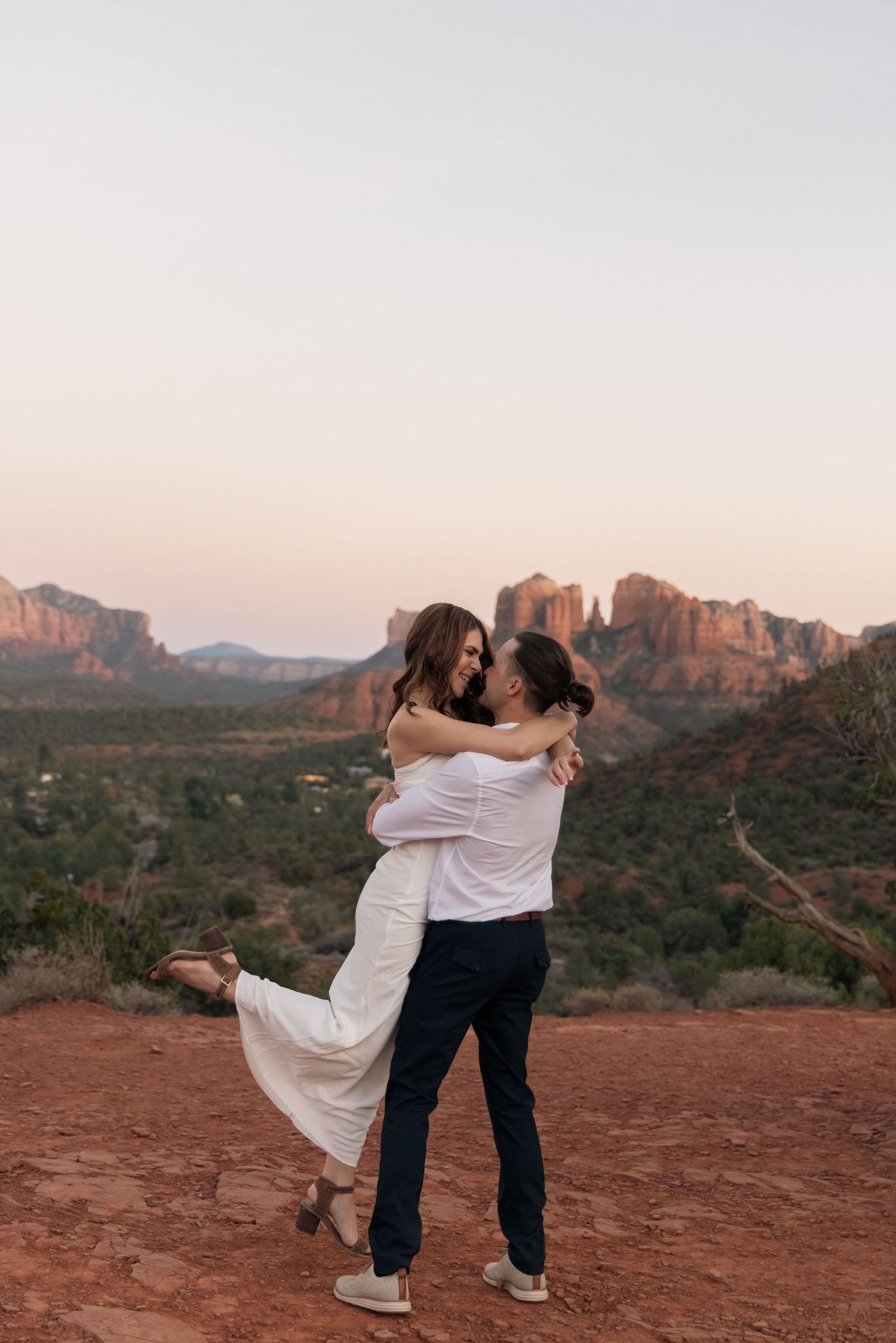 A couple embraces and dances together on red rocks overlooking scenic desert mountains at sunset.