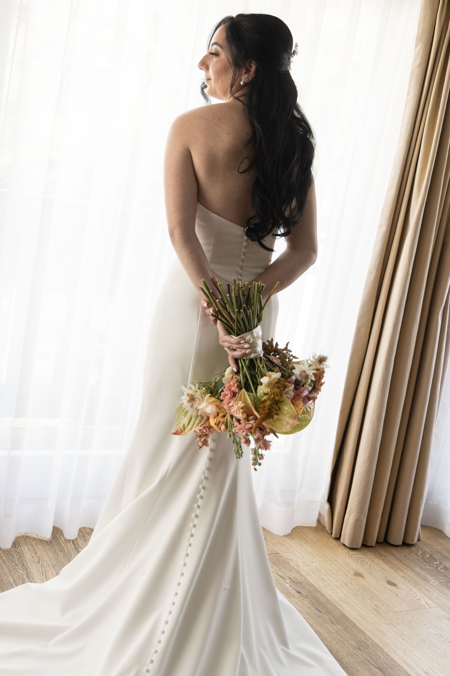 Bride in a strapless satin wedding gown with button back and long train holds a pastel bouquet behind her while gazing out a sunlit window.