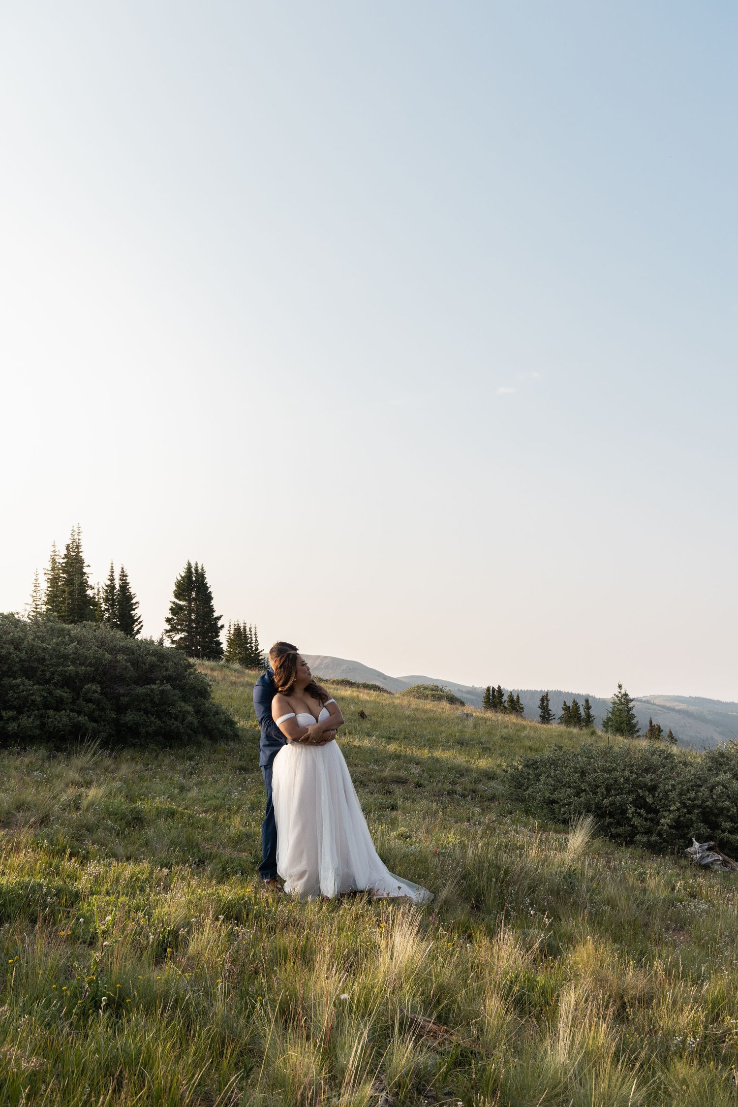Bride and groom embrace on a lush mountain meadow in golden hour light, surrounded by evergreen trees.