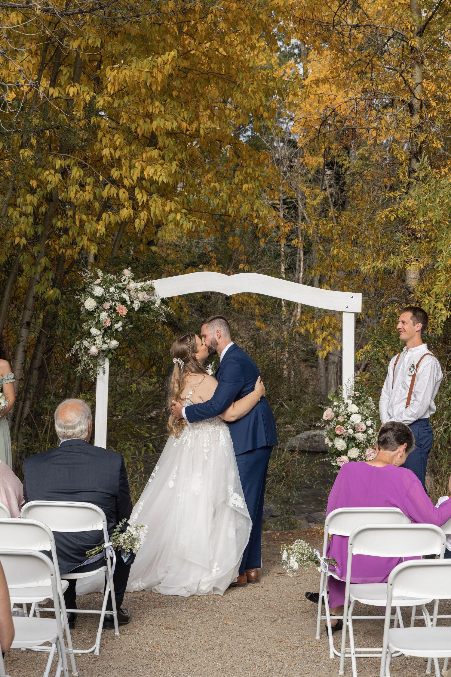 Couple shares first kiss at outdoor fall wedding ceremony under white floral arch with golden autumn trees backdrop.