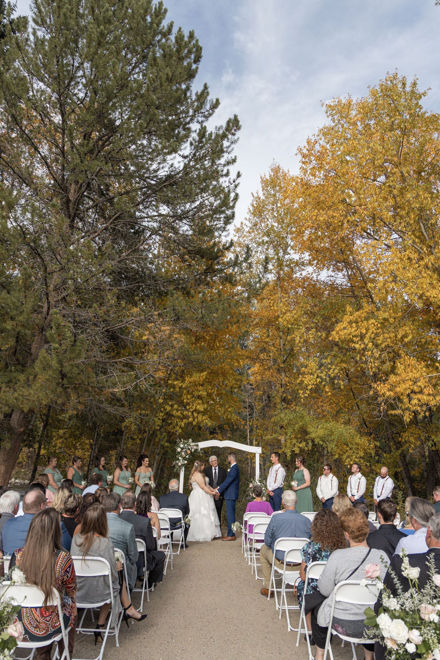 Outdoor fall wedding ceremony with guests seated in white chairs, couple at altar surrounded by colorful autumn trees.