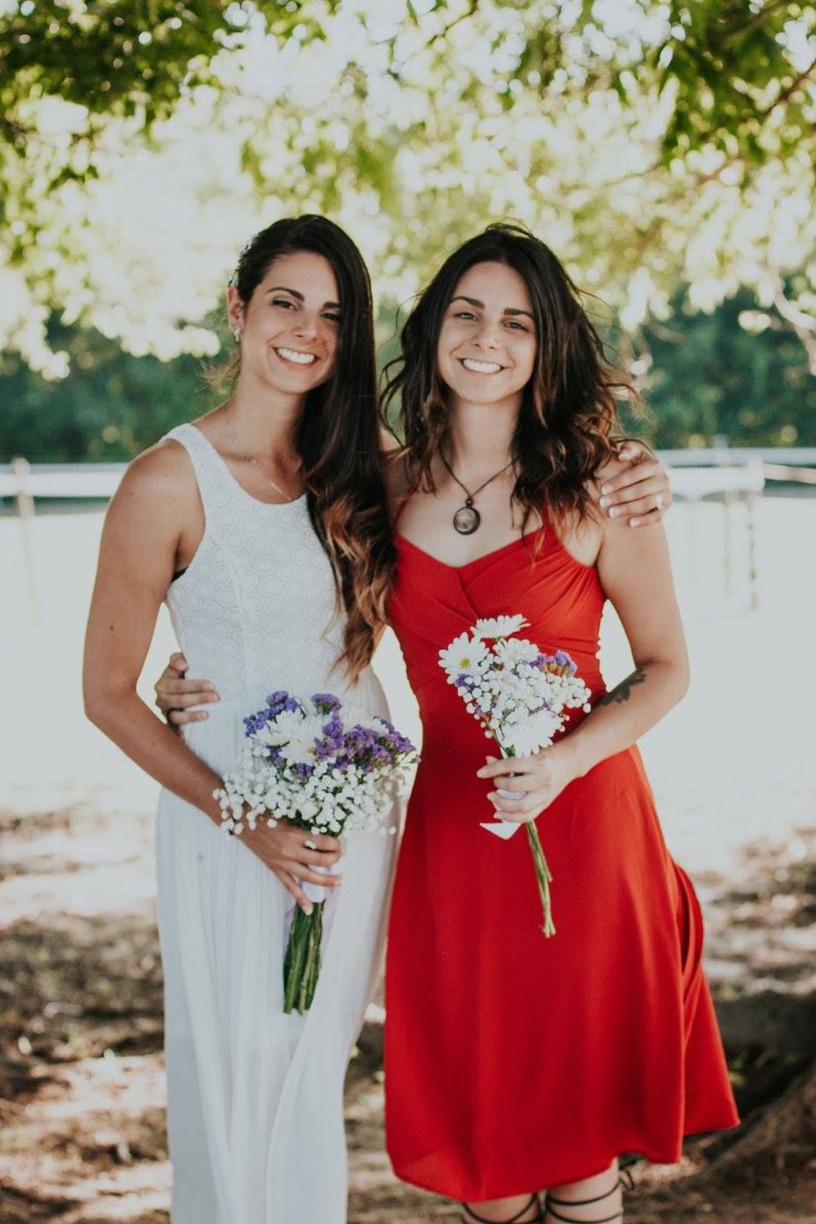 Two smiling women in white and red dresses holding flower bouquets outdoors at a wedding celebration.