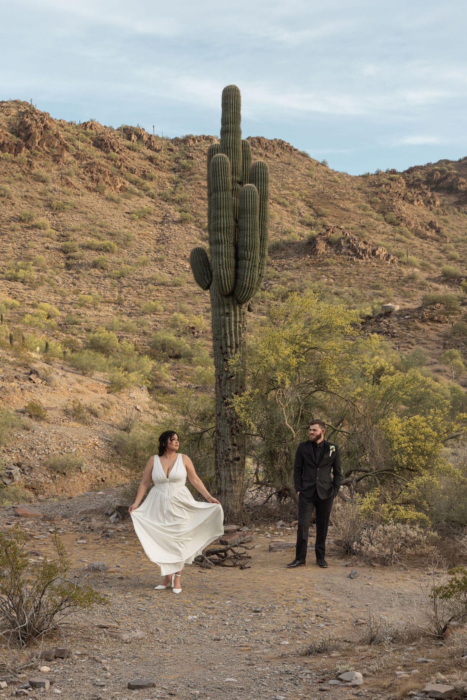 A couple poses for wedding photos in a desert landscape with tall saguaro cactus and rocky mountains in the background.