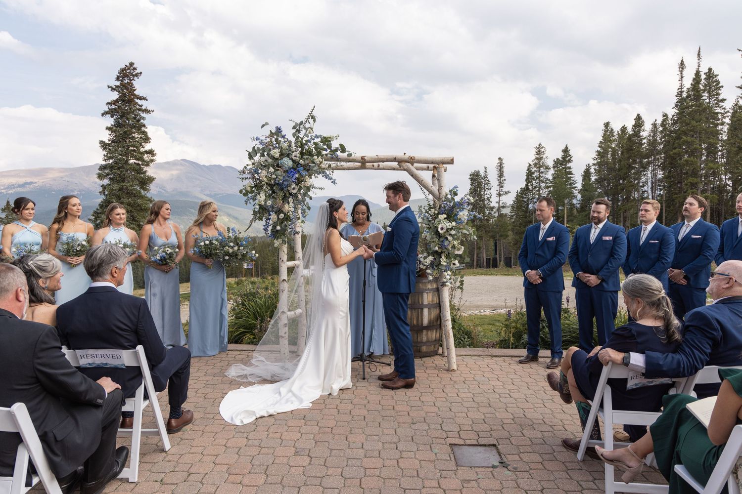 Outdoor mountain wedding ceremony with bride and groom at floral arch, bridesmaids in blue, groomsmen in navy suits.