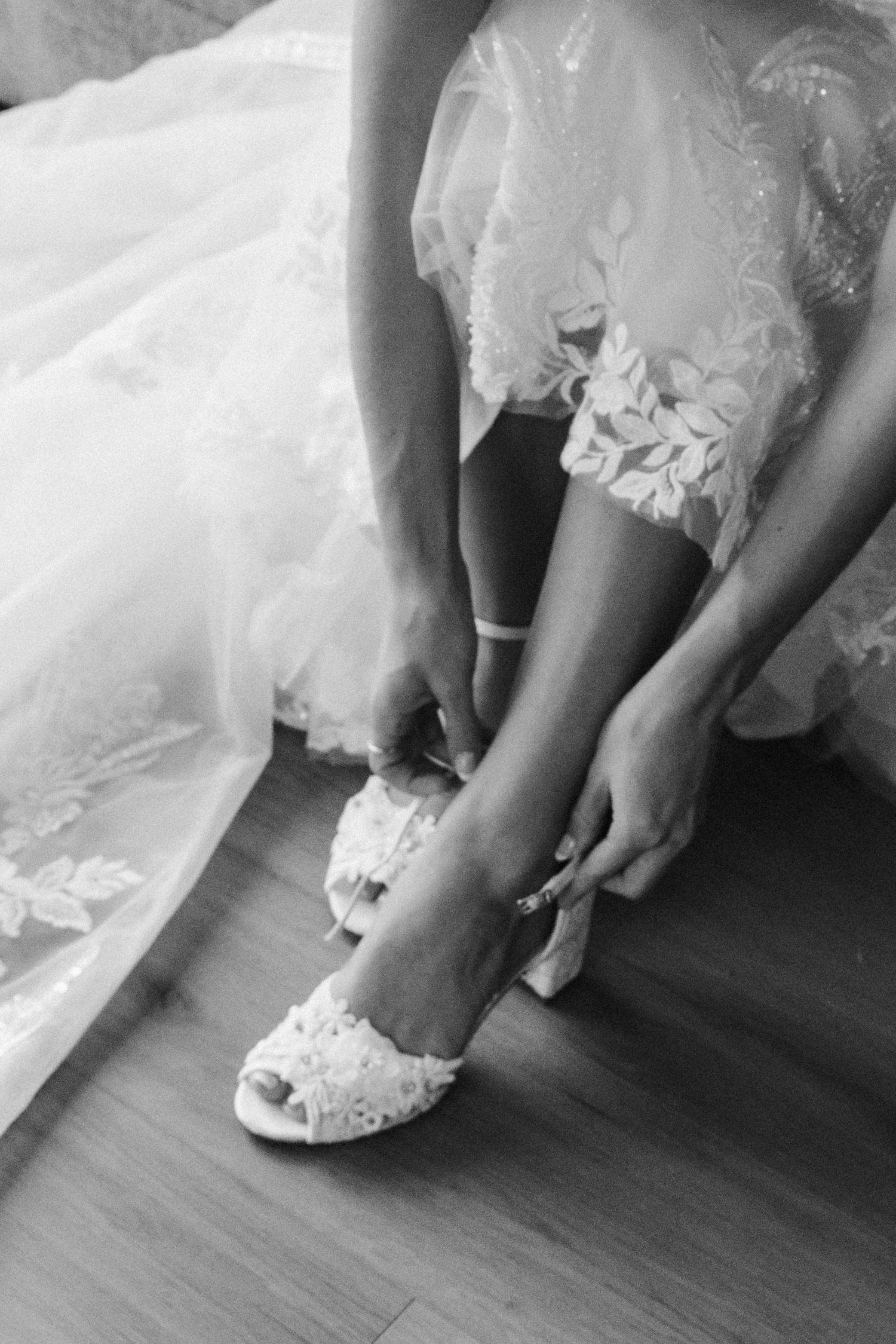 Bride putting on lace wedding heels in black and white photo, adjusting ankle strap of bridal shoe.