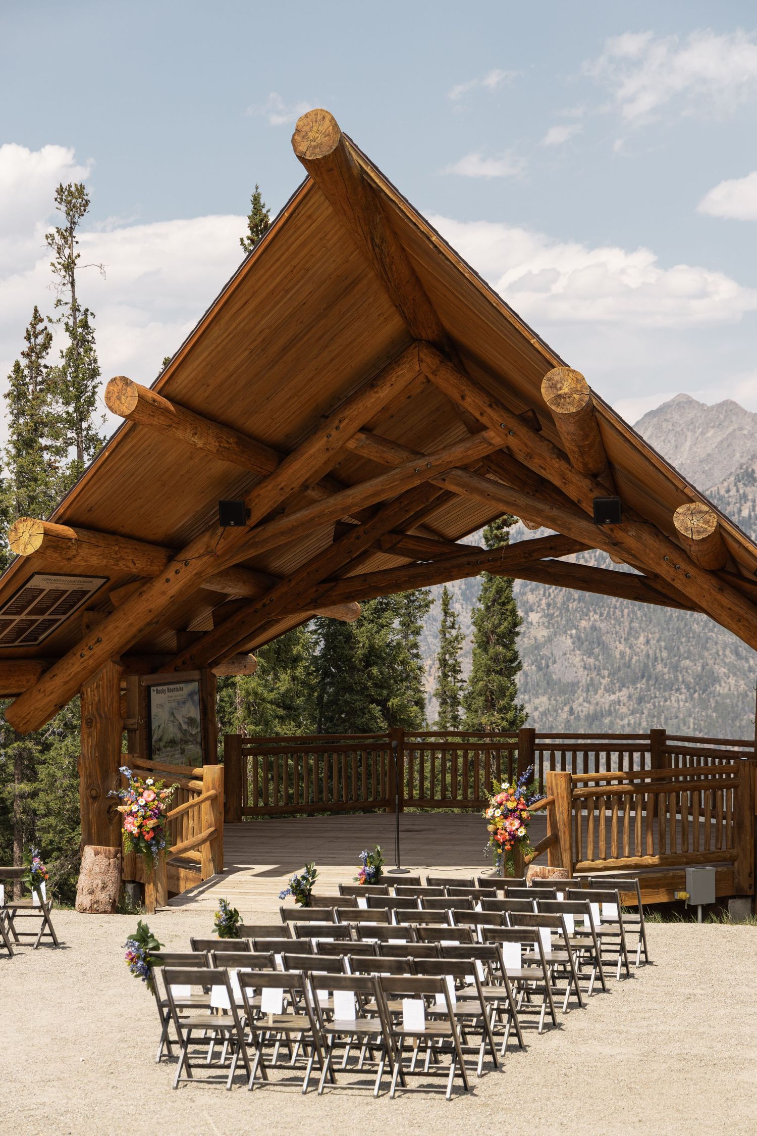 Rustic log pavilion wedding ceremony setup with mountain views, rows of chairs, and floral arrangements in a scenic outdoor venue.