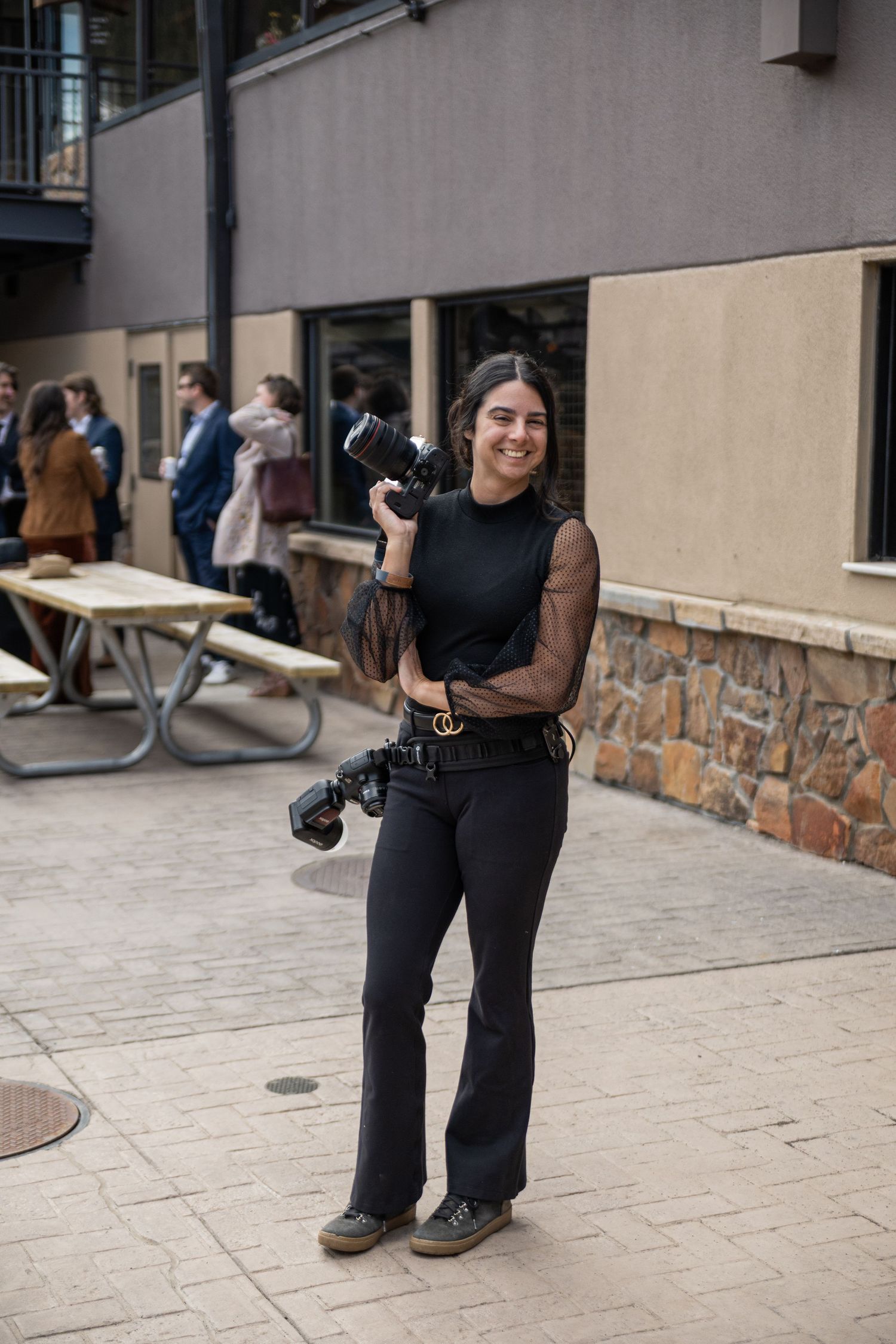 Smiling woman in black outfit holding a professional camera outdoors on a stone patio near a building.