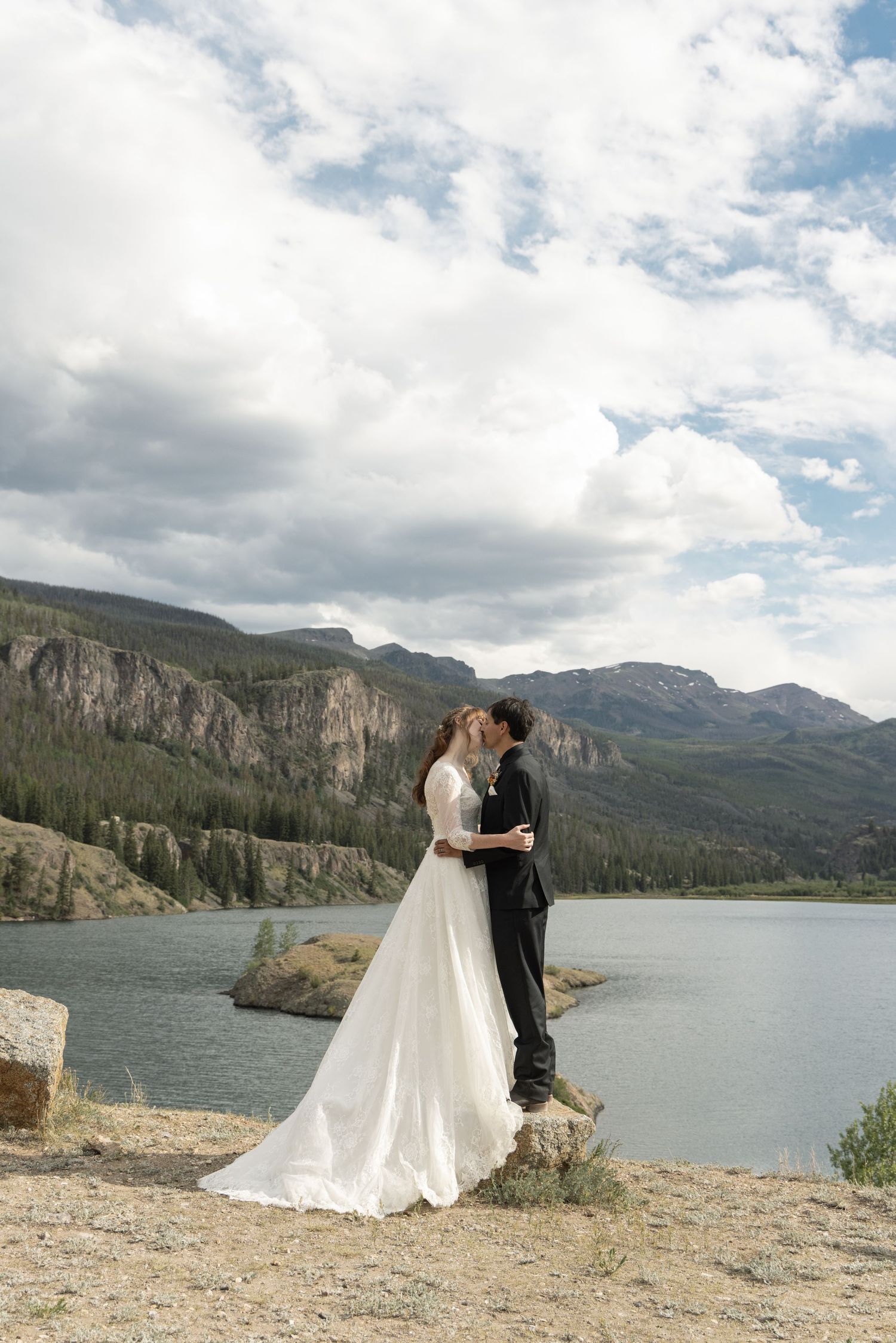 Bride and groom kissing on rocky lakeside with mountain backdrop under cloudy sky in scenic outdoor wedding photo.