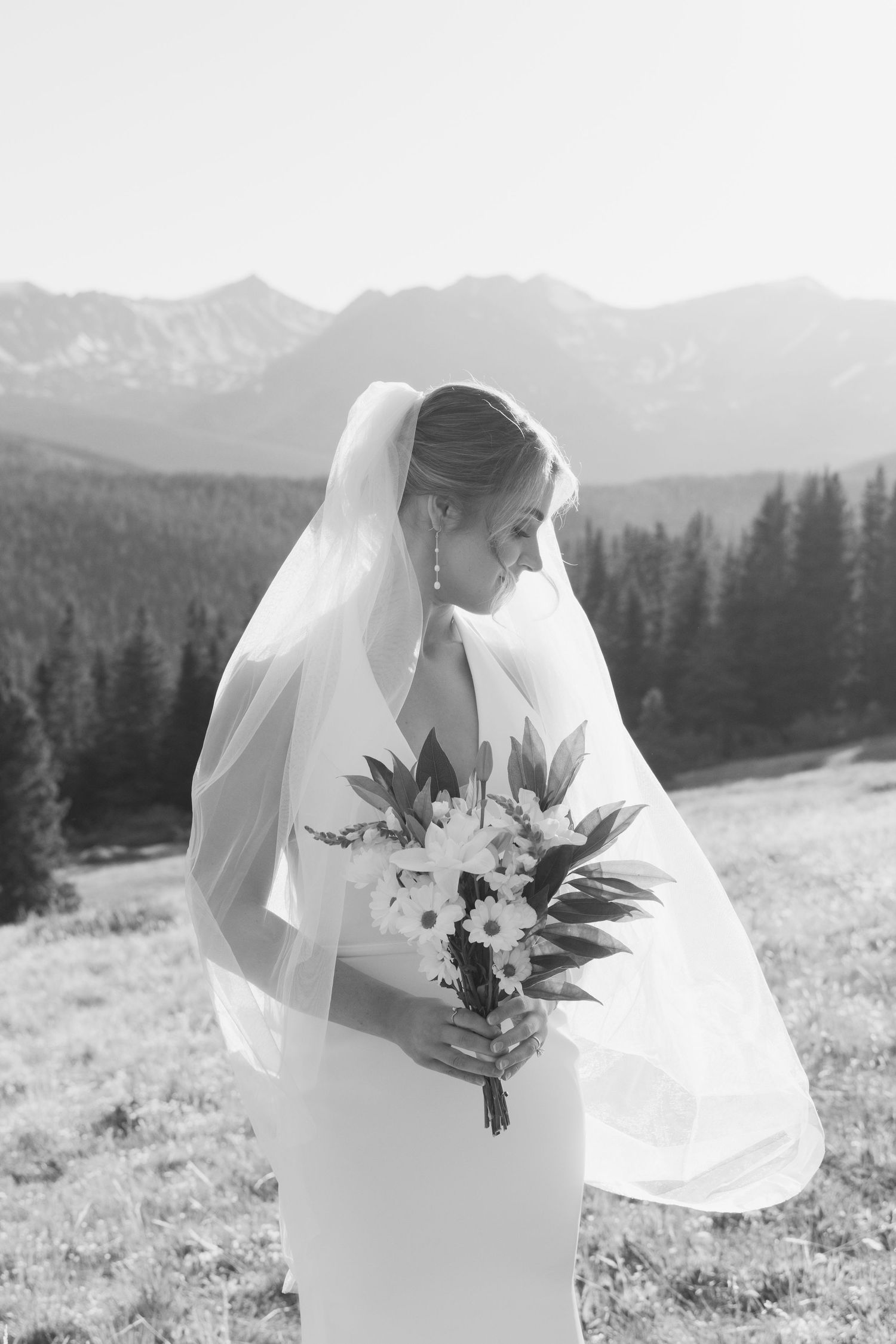 Black and white photo of a bride in a veil holding a bouquet with mountain and forest backdrop.
