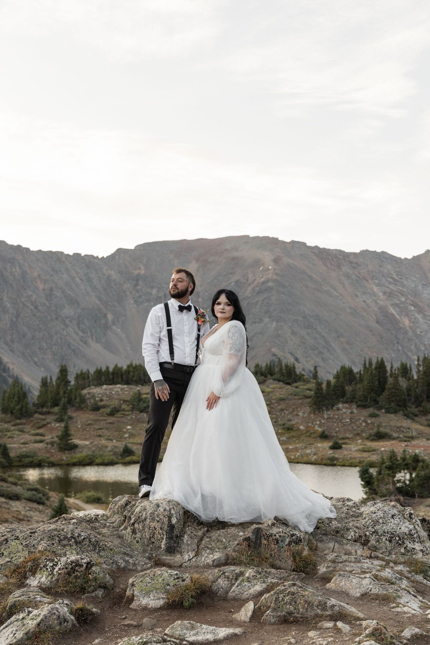 Bride and groom pose on rocky mountain landscape near alpine lake, she in white gown, he in suspenders and bow tie.