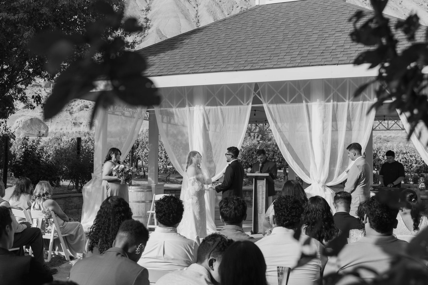 Black and white outdoor wedding ceremony under draped gazebo with guests seated watching couple exchange vows.