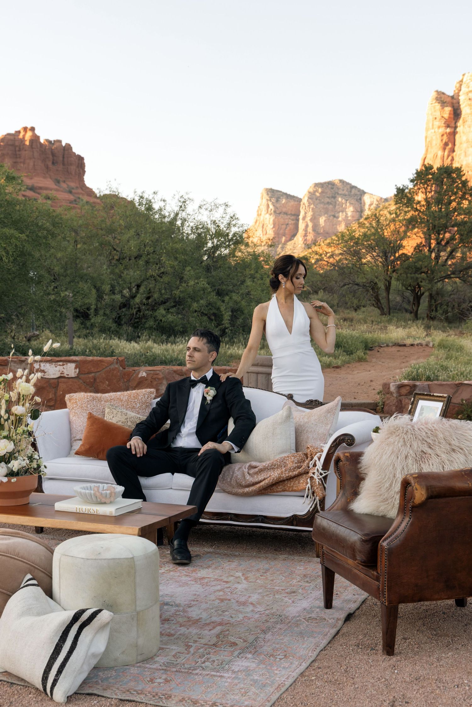 A bride and groom pose on elegant outdoor lounge furniture with stunning Sedona red rock formations at sunset.
