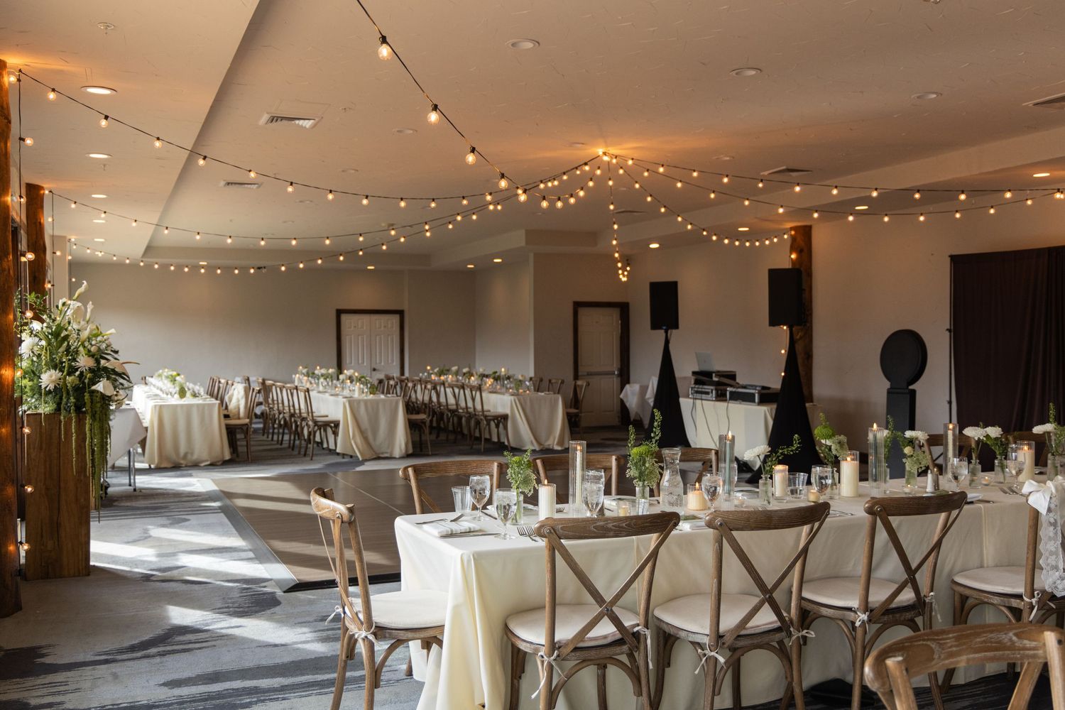 Elegant wedding reception hall with string lights, round tables, cross-back chairs, and floral centerpieces in a modern venue.
