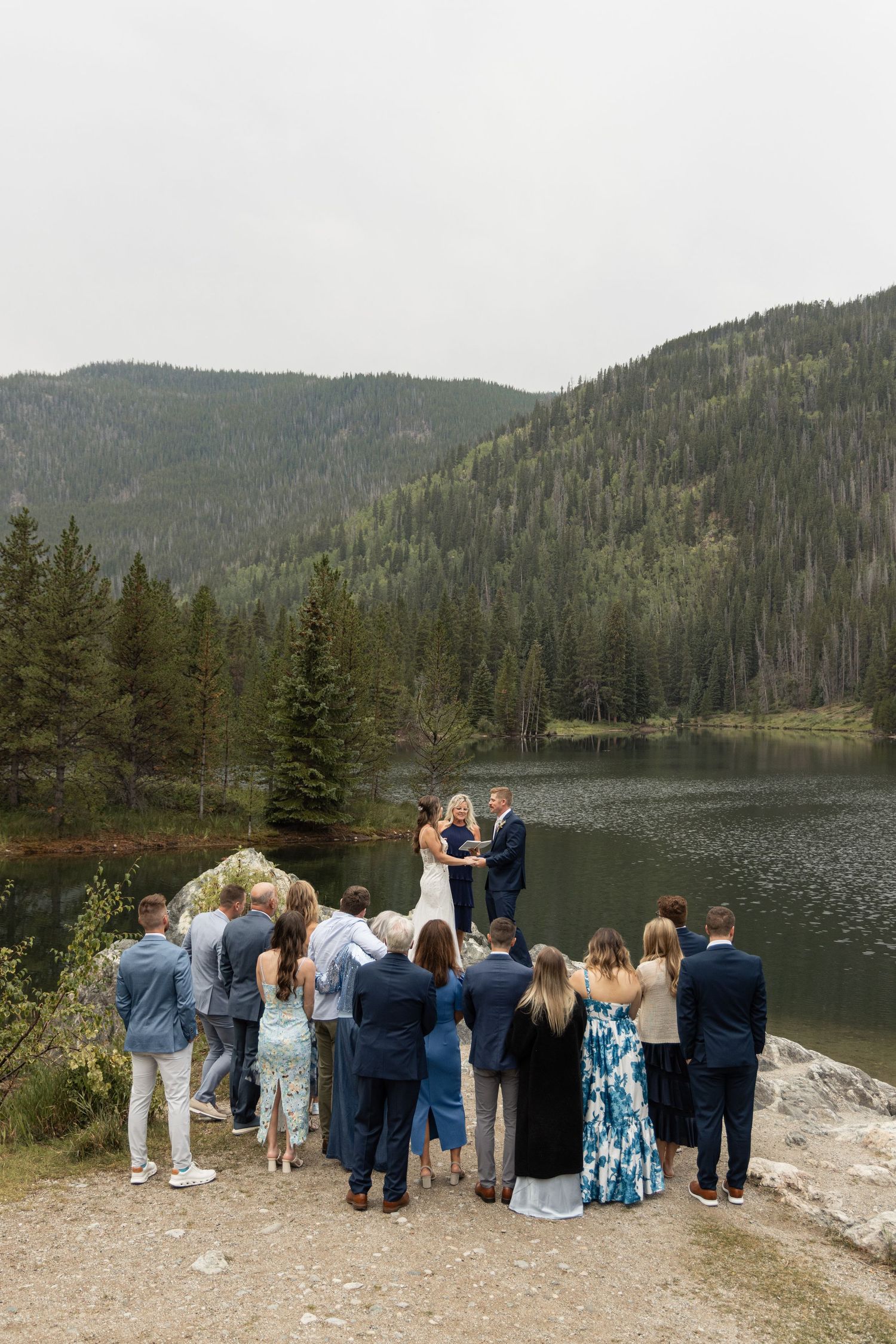 Outdoor wedding ceremony by a mountain lake surrounded by pine trees, with guests gathered around the couple.