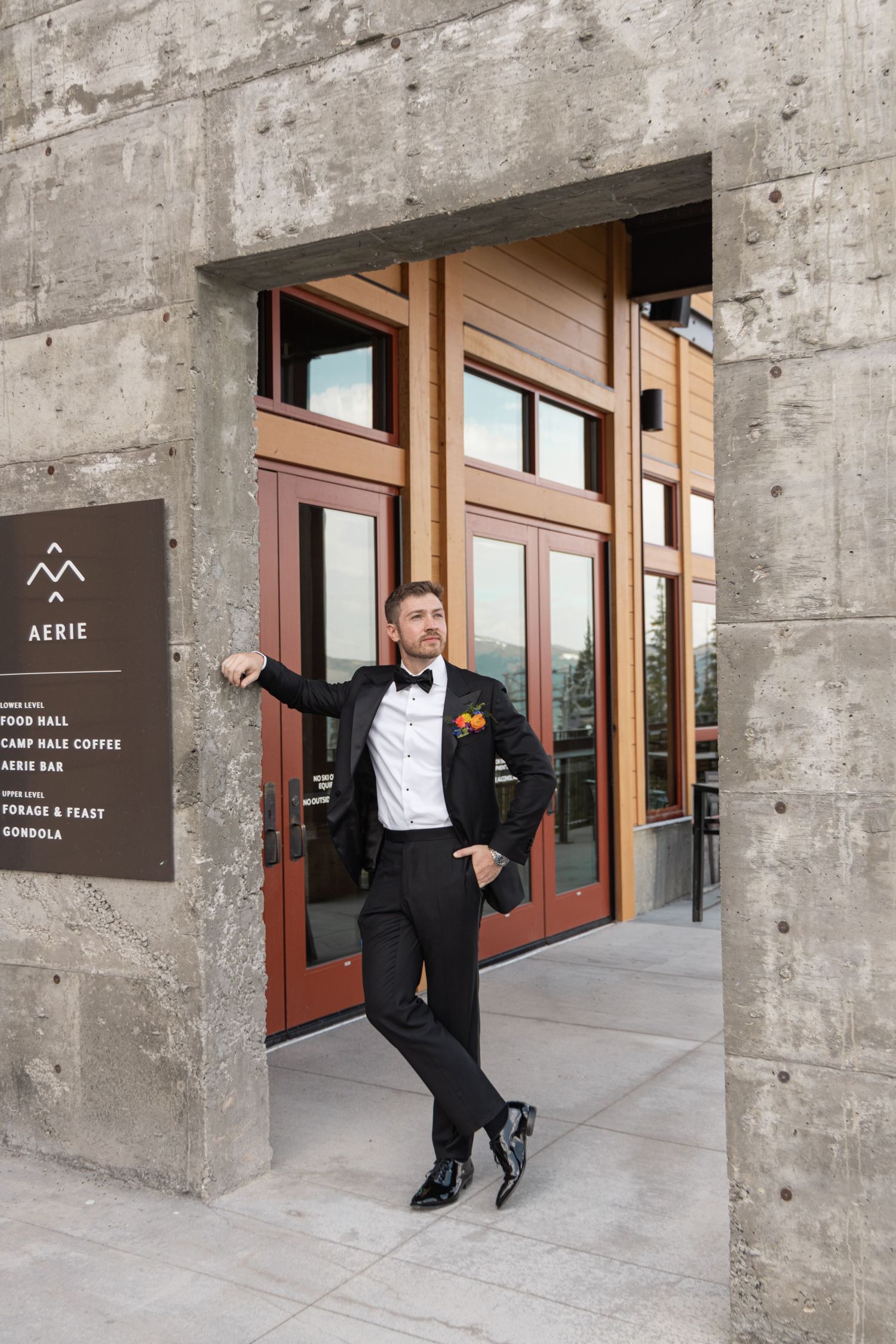 Groom in black tuxedo with bow tie poses at Aerie venue entrance, leaning against stone wall with wooden doors.