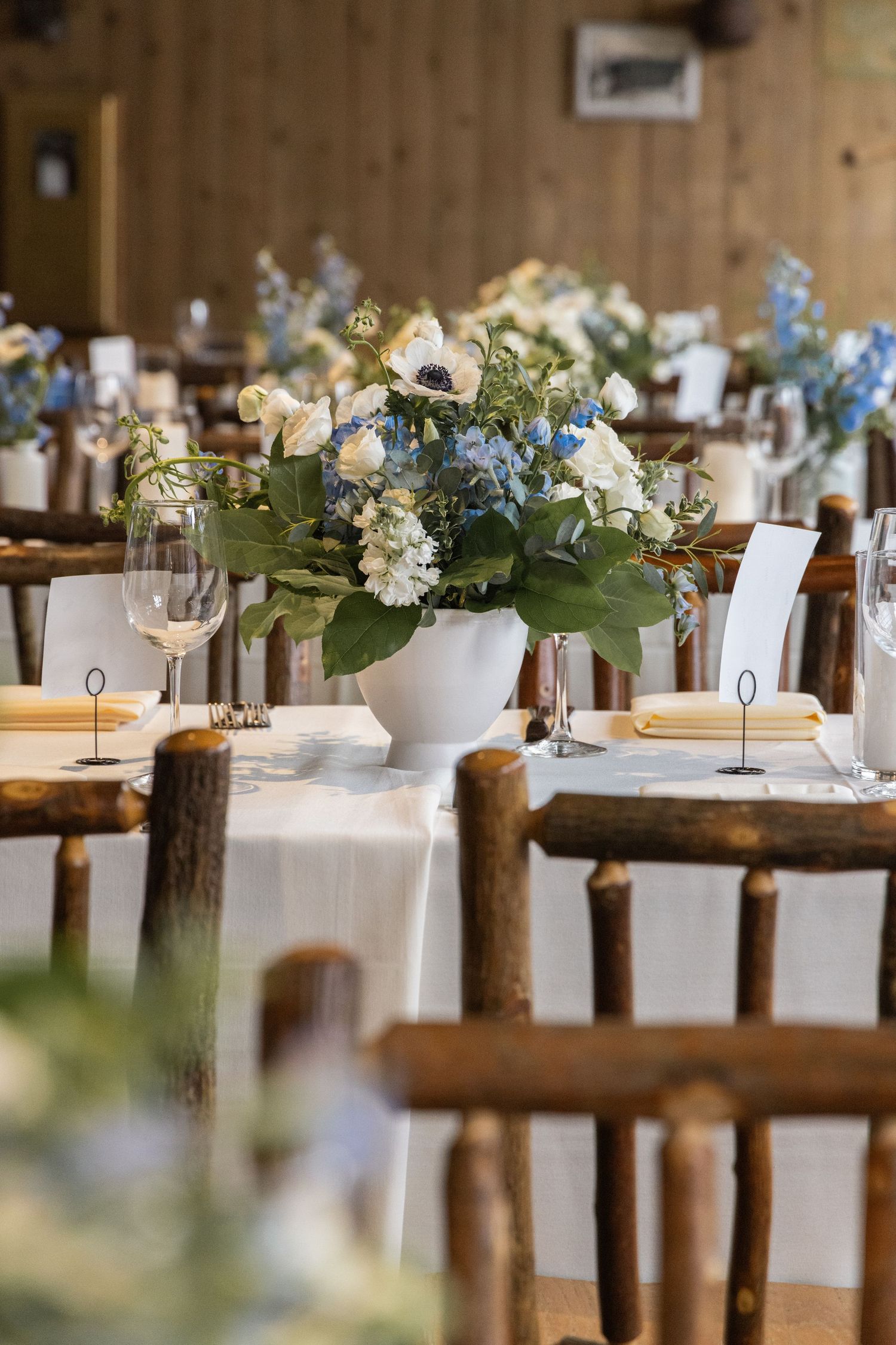 Rustic wedding reception table with white floral centerpiece featuring blue and white blooms, wooden chairs, and elegant place settings.