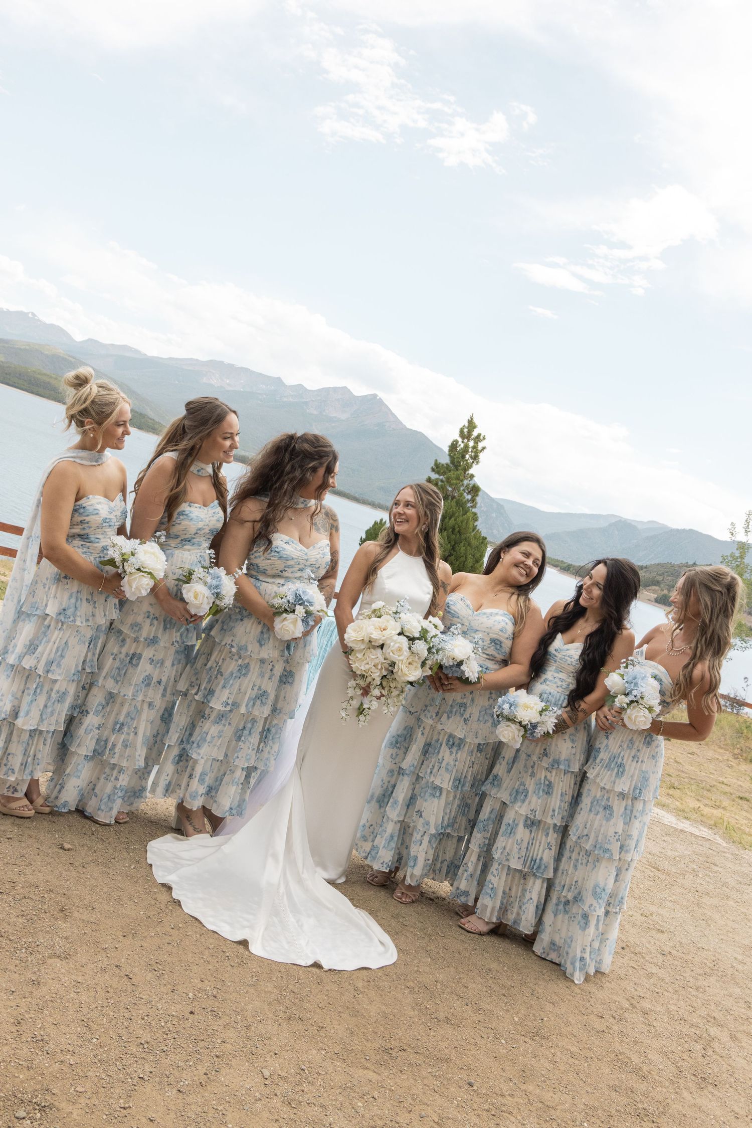 Bride in white gown with bridesmaids in blue floral tiered dresses holding bouquets near mountain lake.