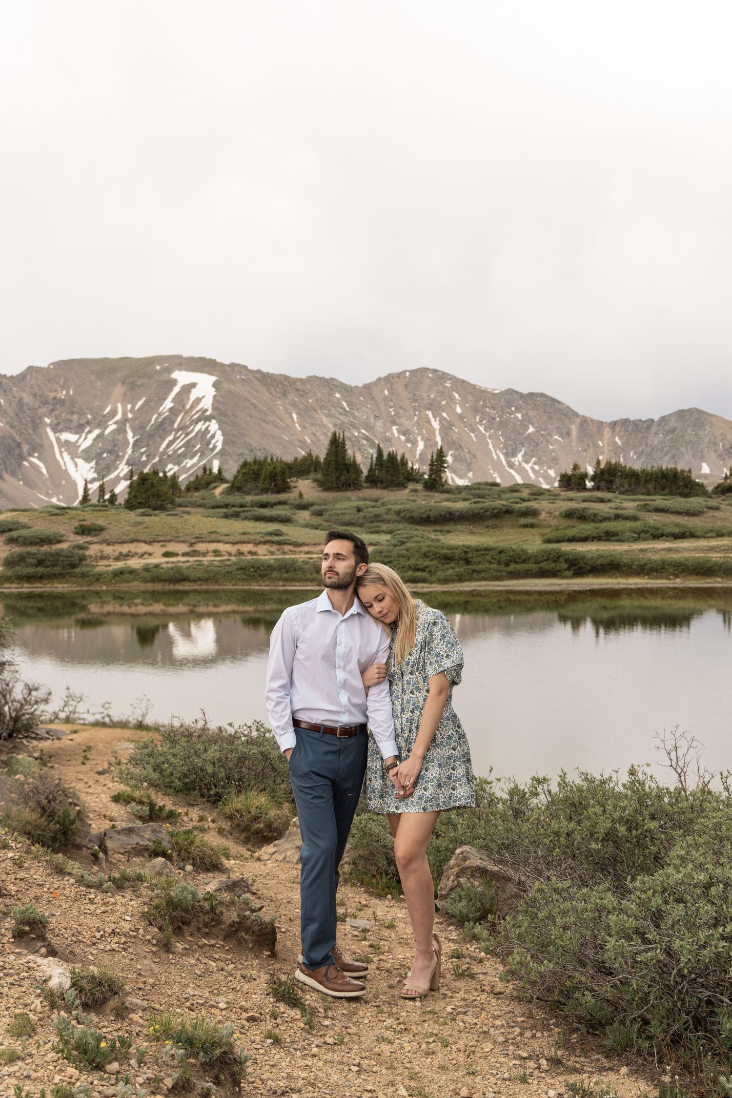 A couple shares a romantic moment by a serene mountain lake with snow-capped peaks in the background.