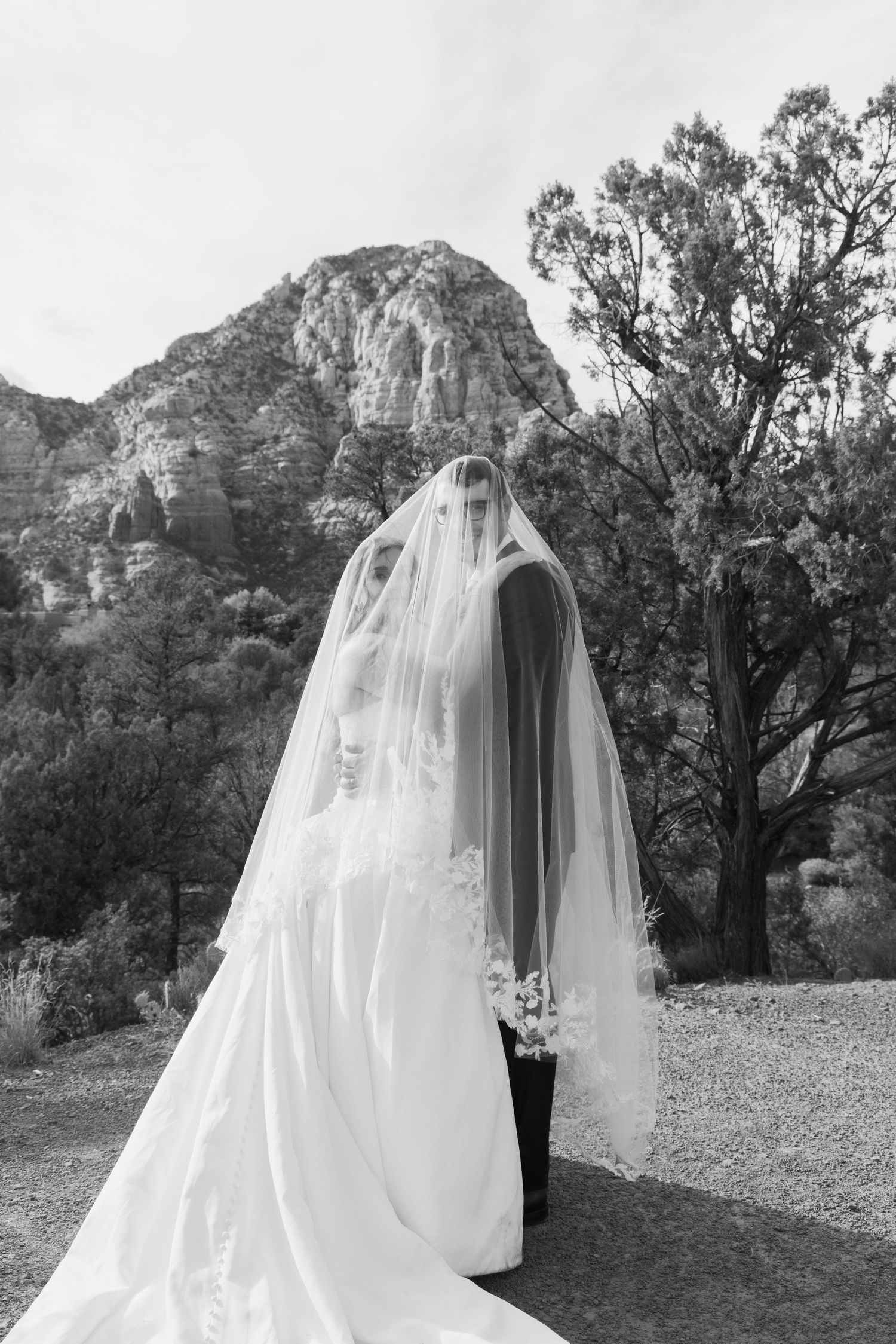 Black and white photo of bride and groom kissing under flowing veil with rocky mountain landscape backdrop.