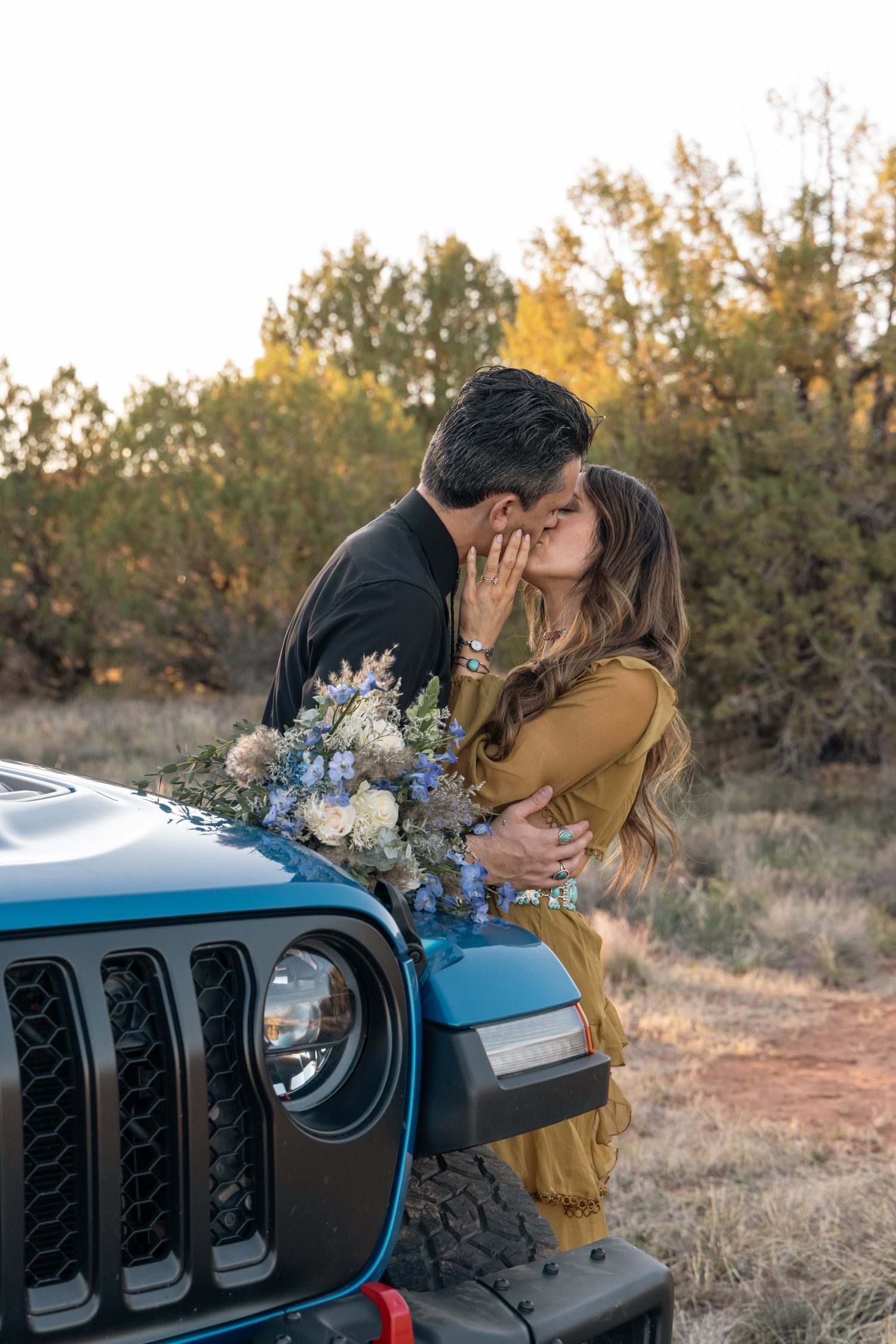 Romantic couple shares a tender kiss while leaning against a blue Jeep with flowers in an autumn desert setting.