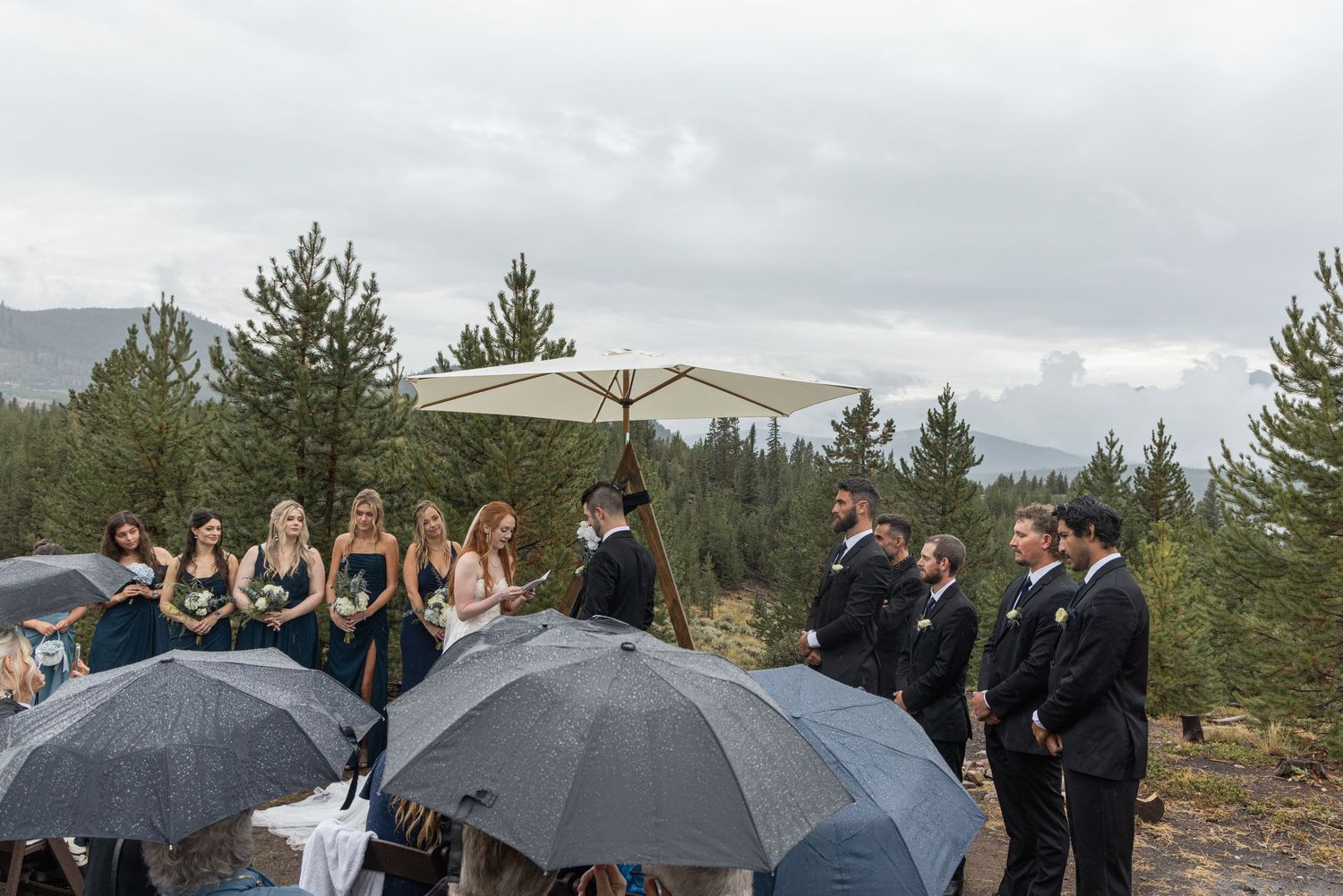 Wedding party stands together under black umbrellas during a rainy outdoor mountain ceremony.