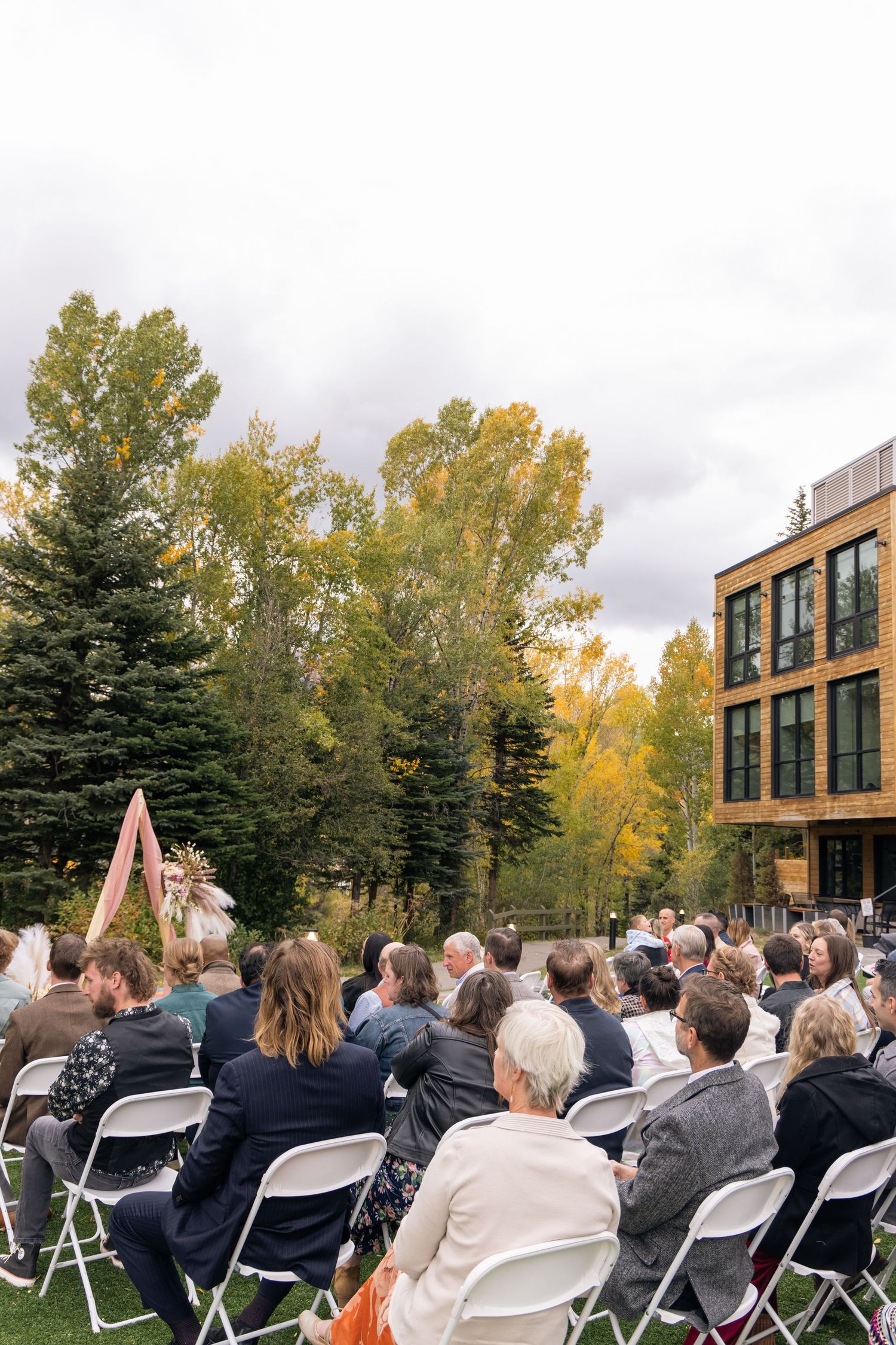 Outdoor wedding ceremony with guests seated on white chairs, a floral arch, autumn trees, and a modern building backdrop.