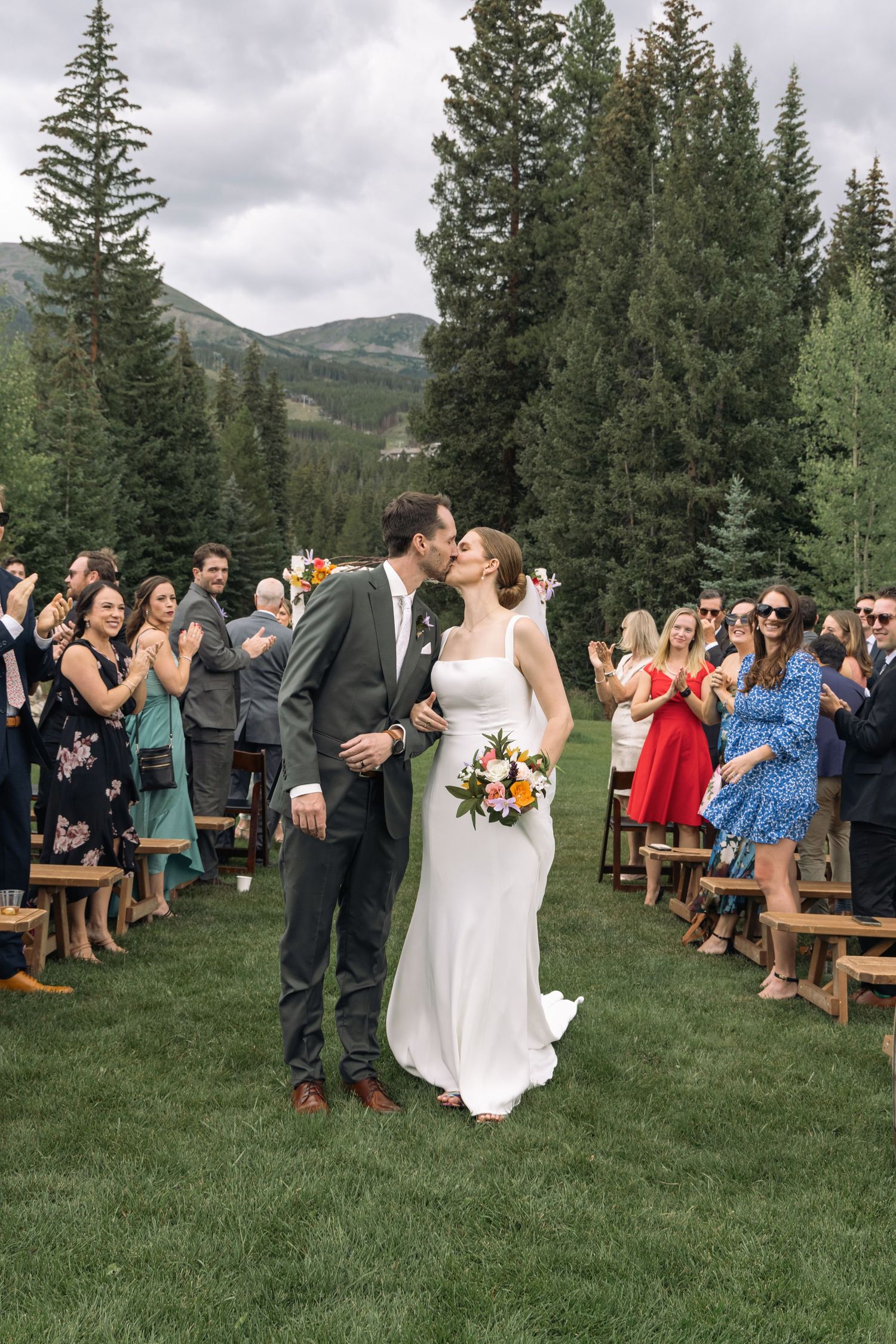Bride and groom share a kiss at outdoor mountain wedding ceremony while guests applaud along the aisle.