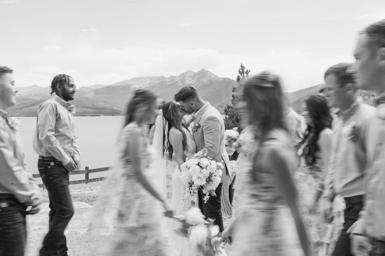 Black and white wedding kiss photo with mountain lake backdrop, guests walking past blurred in foreground.