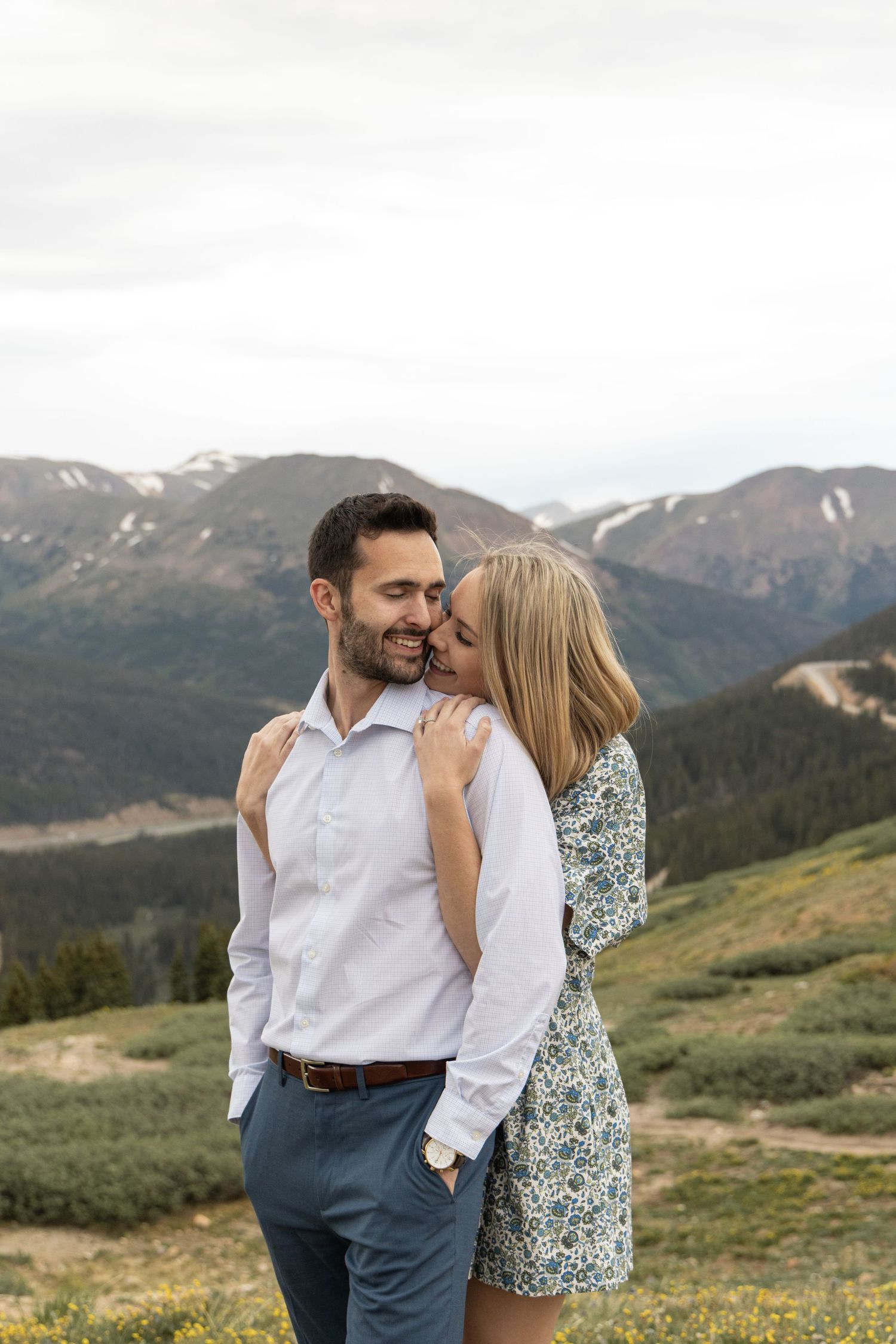 A romantic couple shares an intimate moment while standing on a mountain meadow with snow-capped peaks in the background.