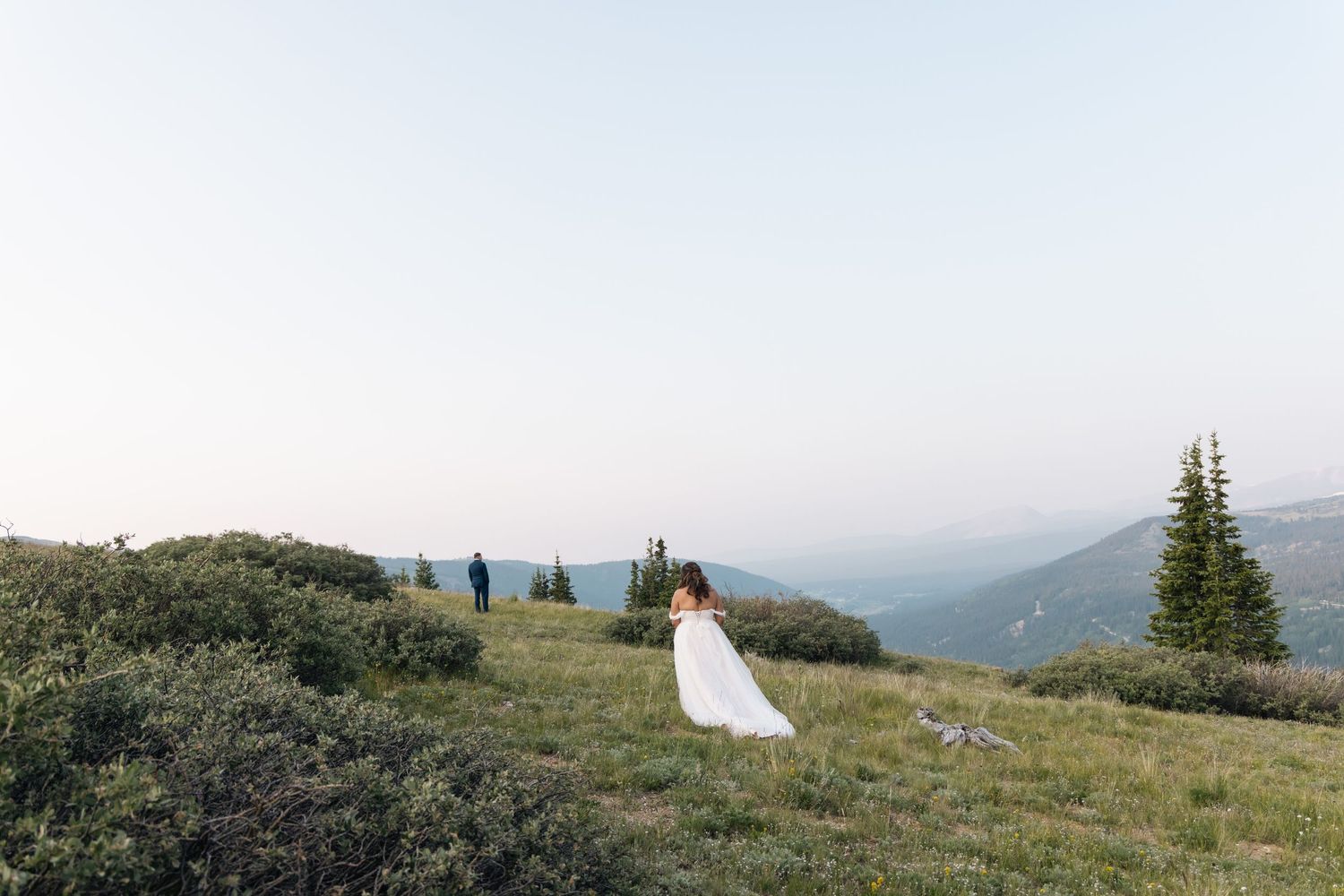 Bride in white gown stands on a lush mountain hillside with sweeping valley views during an outdoor wedding ceremony.