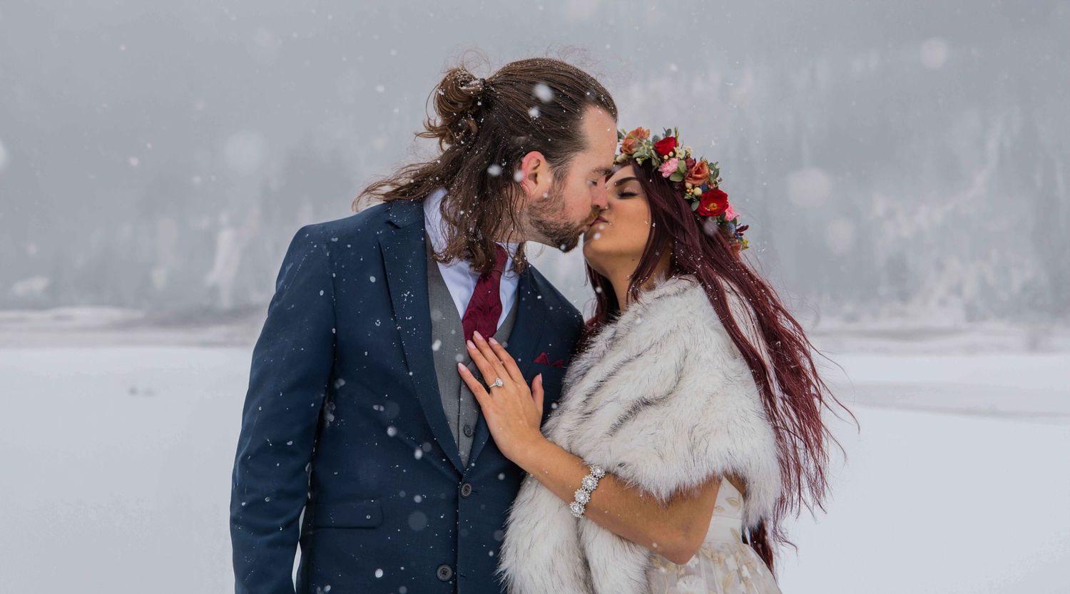 A couple shares a romantic kiss in the snow while wearing winter coats during a snowy photoshoot.
