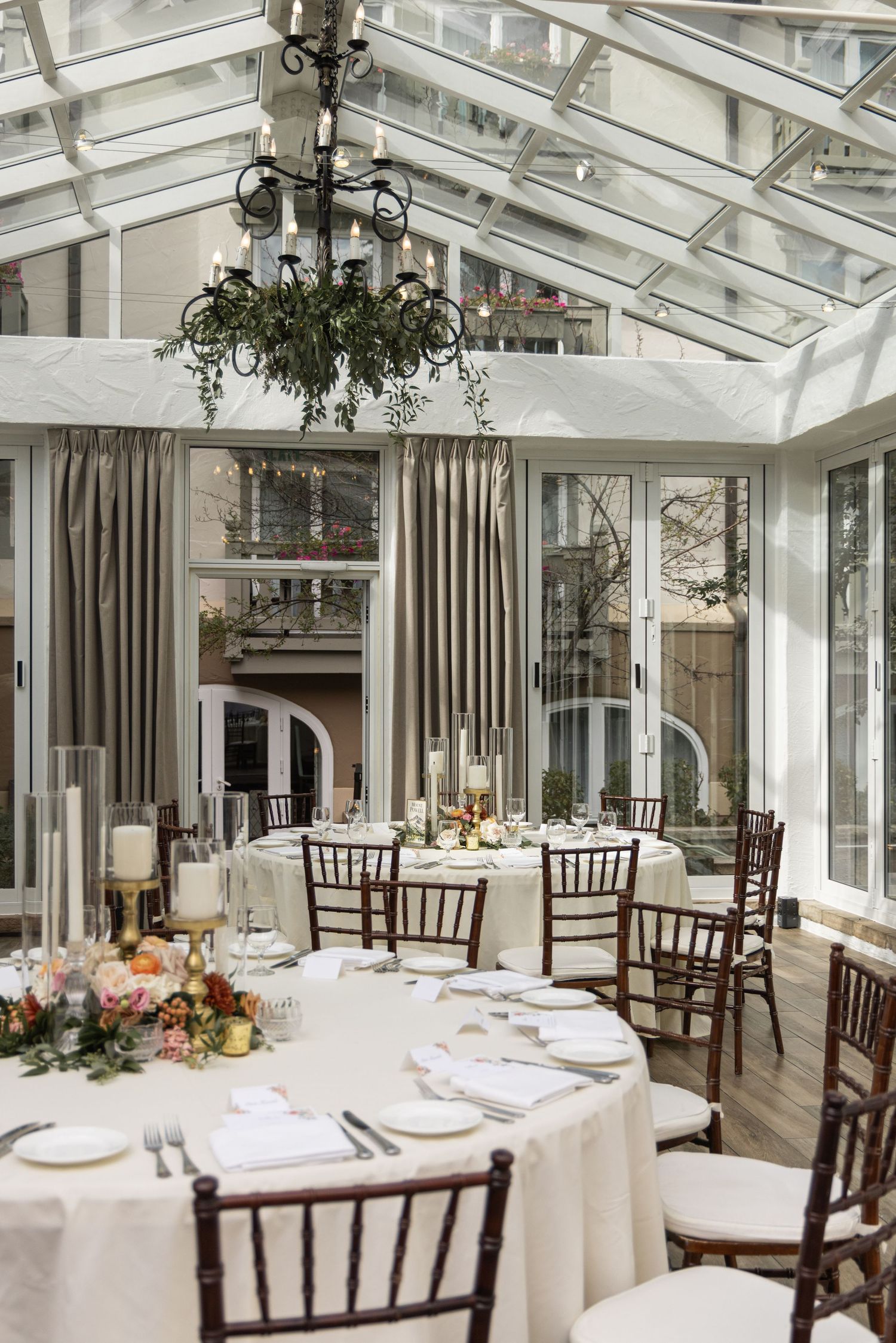 Elegant conservatory wedding reception with white draped tables, chiavari chairs, floral centerpieces, and glass ceiling.