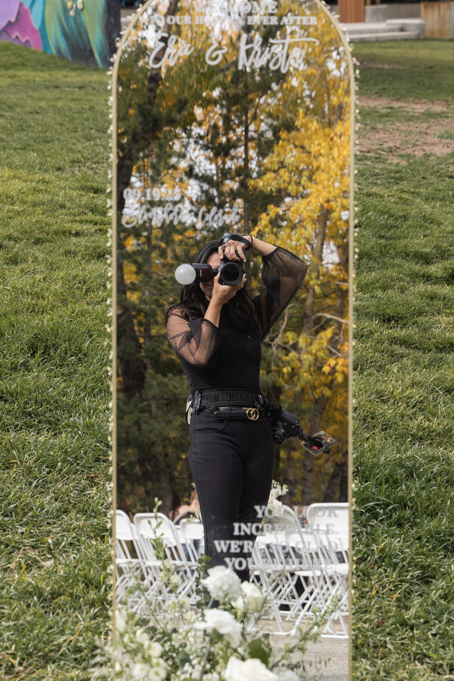 Photographer in black outfit captures reflection in gold mirror wedding sign on green grass lawn.