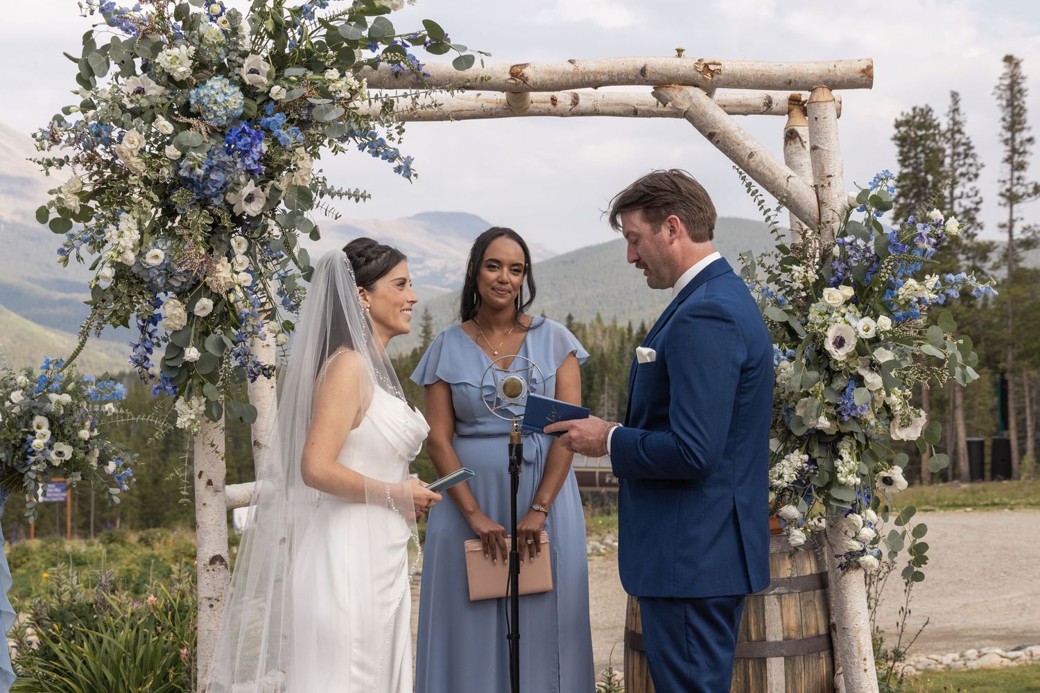 Outdoor mountain wedding ceremony with bride in white gown, groom in navy suit, and officiant under floral arch.