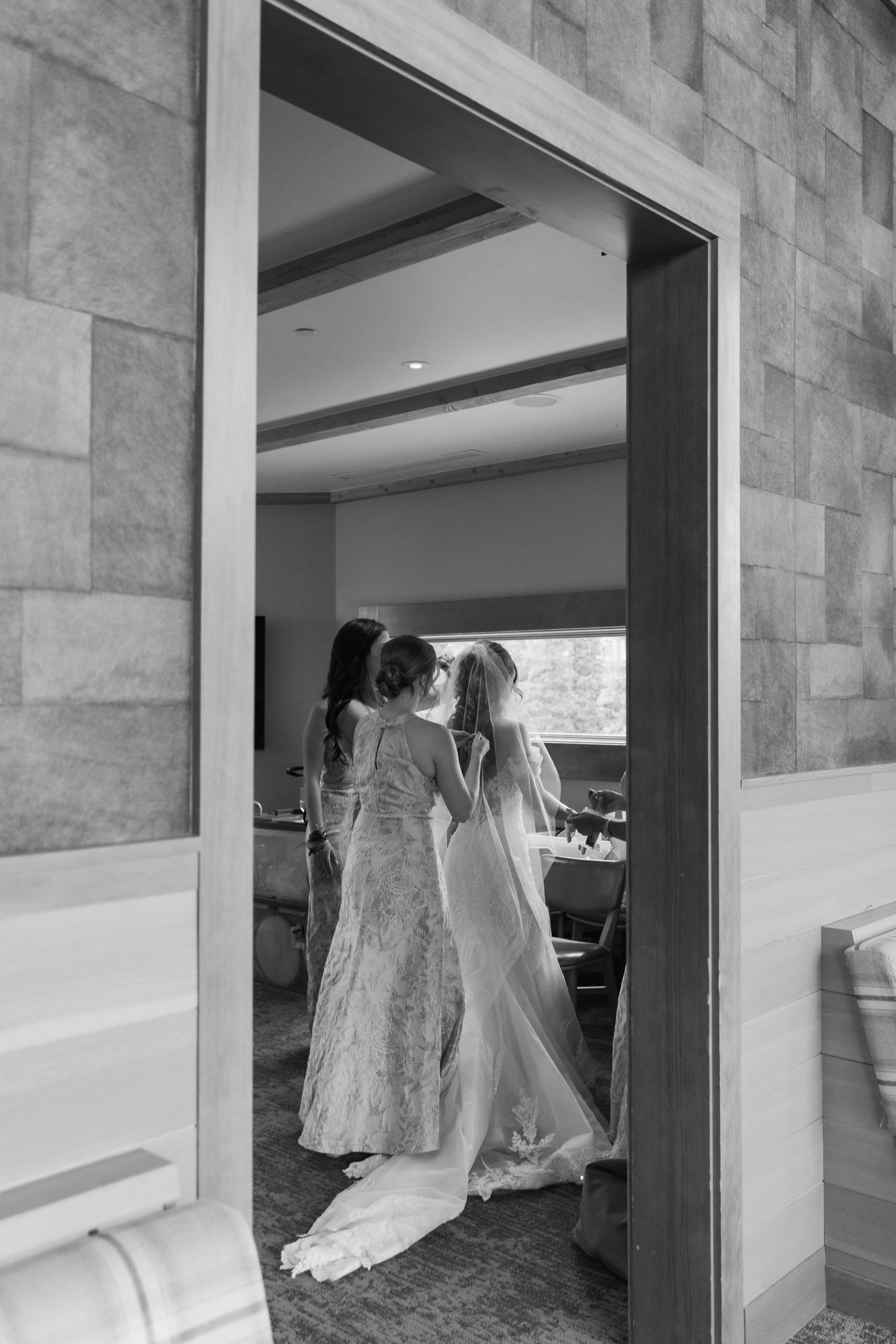Black and white photo of a bride in a flowing gown with attendants helping her prepare in an elegant room.