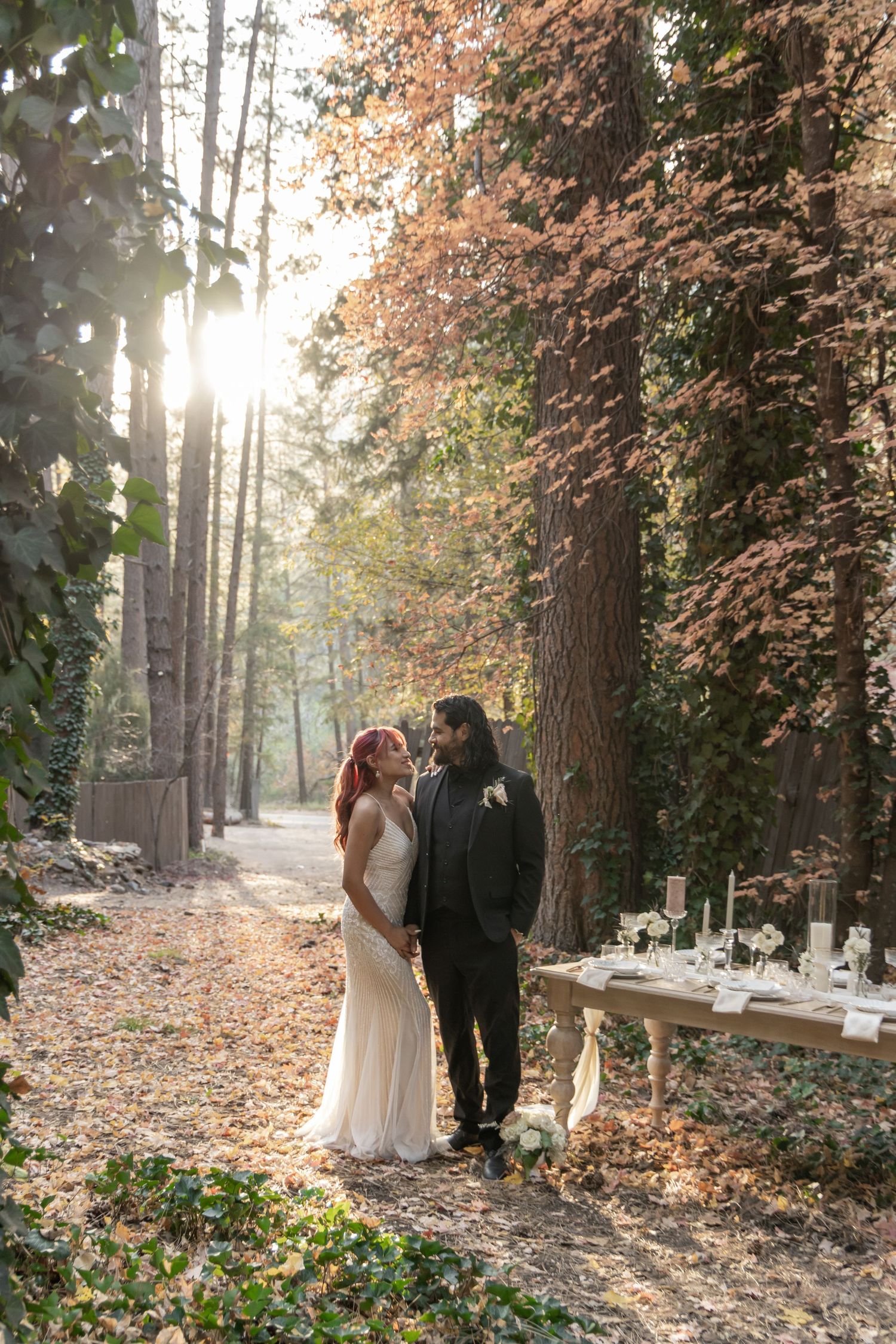 Bride and groom share a romantic moment in a sunlit autumn forest beside an elegant decorated table.