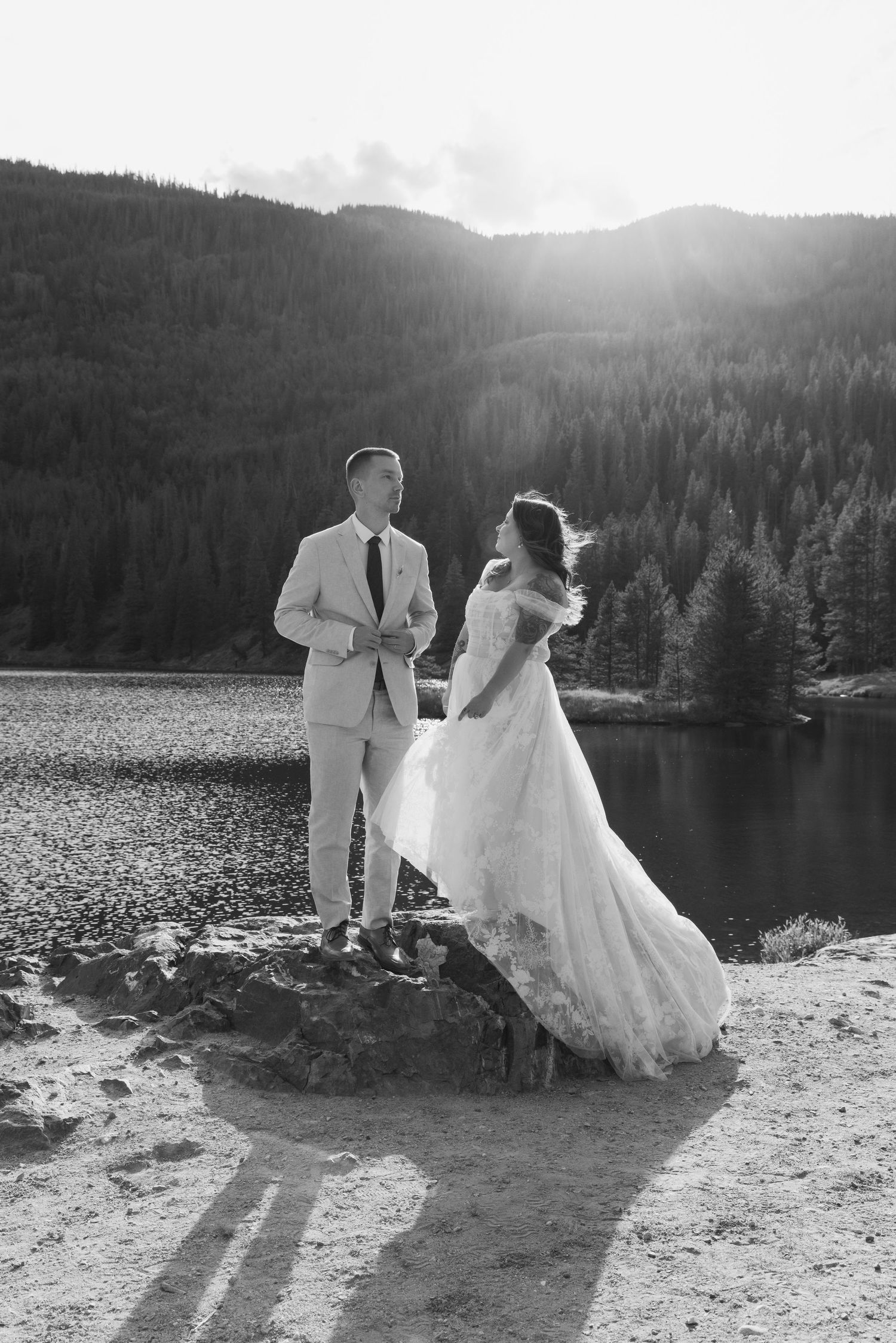 Black and white photo of bride and groom standing on rocks by a mountain lake with forested hills and sunlight behind them.