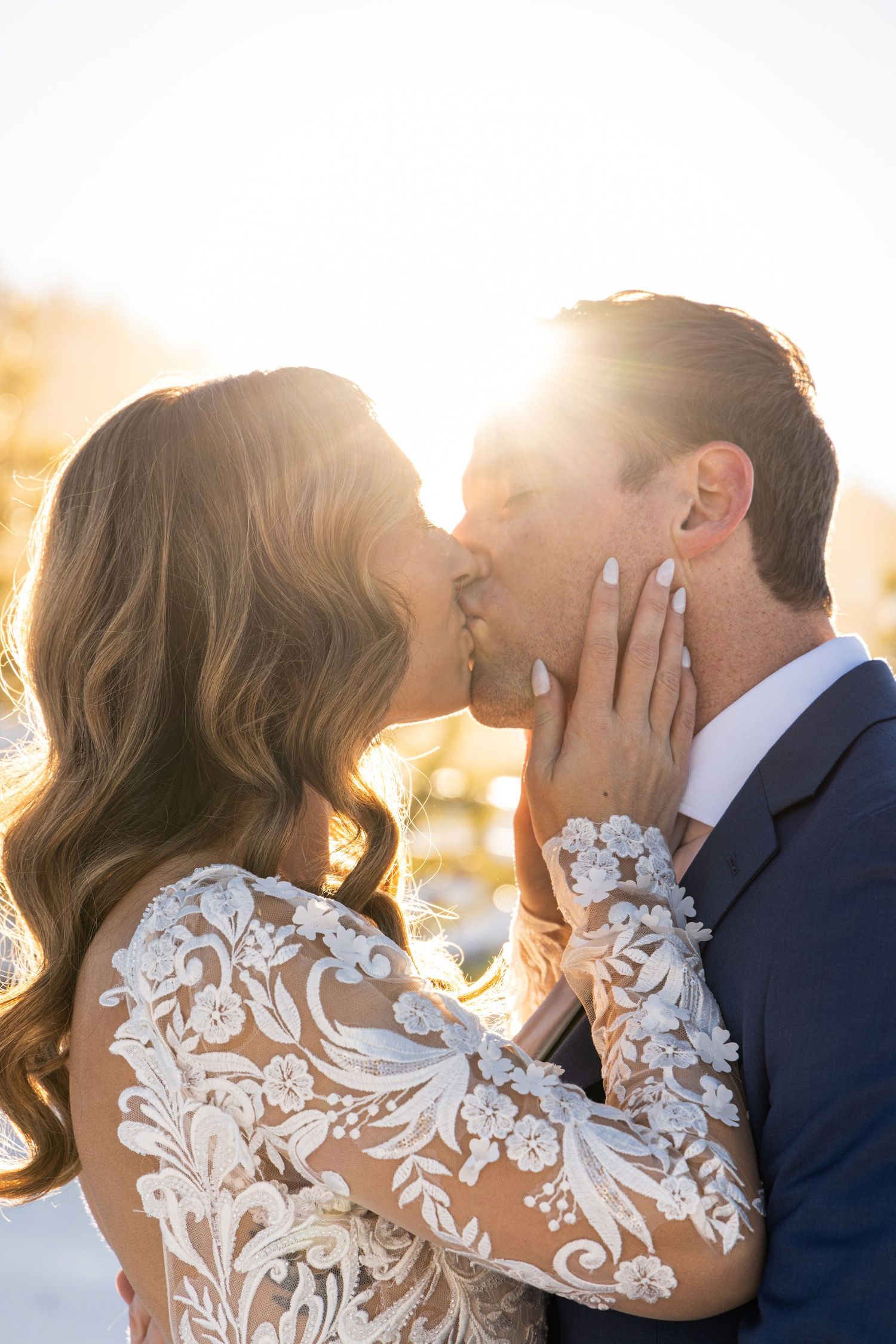 Bride and groom share a romantic golden hour kiss, bride wearing intricate lace long-sleeve wedding gown.