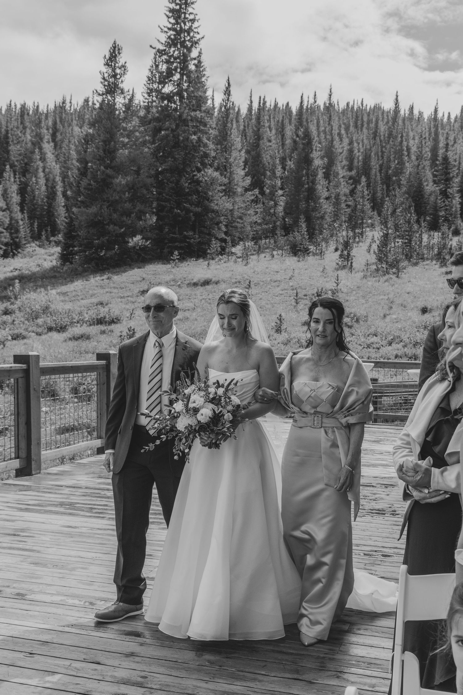 Bride walks down aisle with parents on outdoor wooden deck surrounded by pine trees at mountain wedding ceremony.
